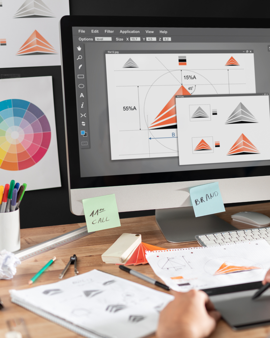 Pantalla de computadora que muestra diagramas y gráficos de diseño de logotipos con formas de pirámides y paletas de colores, en un espacio de trabajo con notas adhesivas y materiales de dibujo.