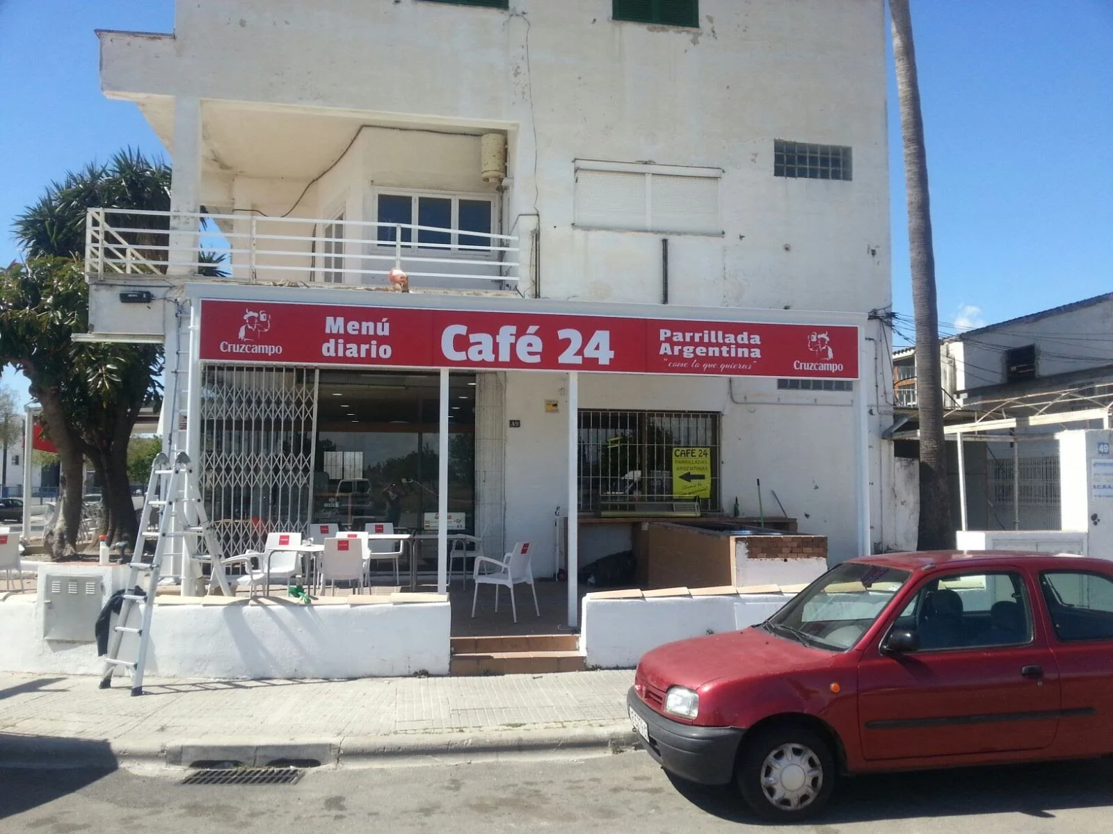 Fachada de un restaurante llamado Café 24 con señalización en color rojo que dice menú diario y parrillada argentina. Hay mesas y sillas afuera, y un automóvil rojo estacionado en la calle.