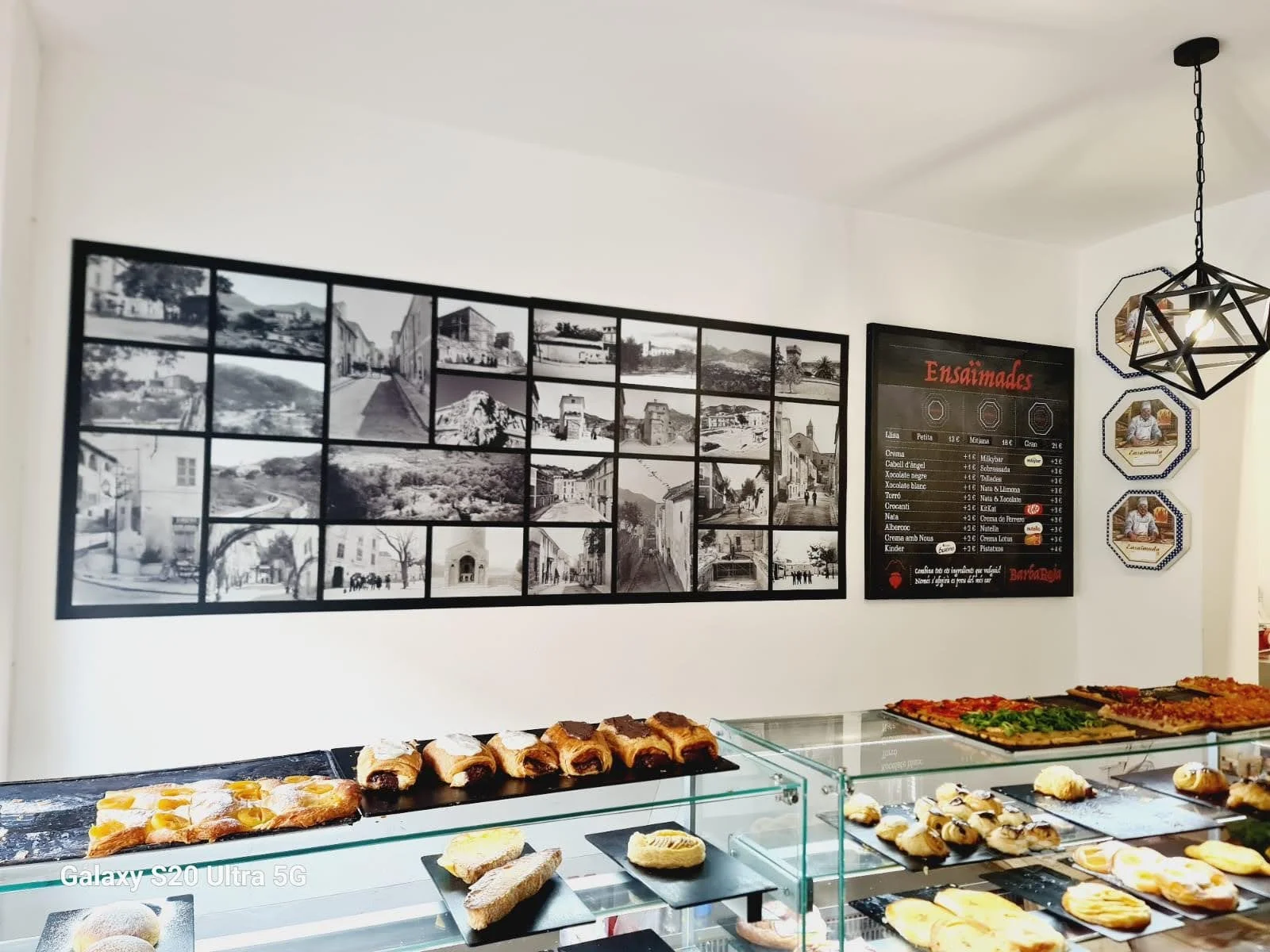 Mostrador con variedad de repostería y pizzas en una panadería o cafetería. En el fondo, cartel con fotos en blanco y negro, y un menú con títulos en rojo y productos en lista