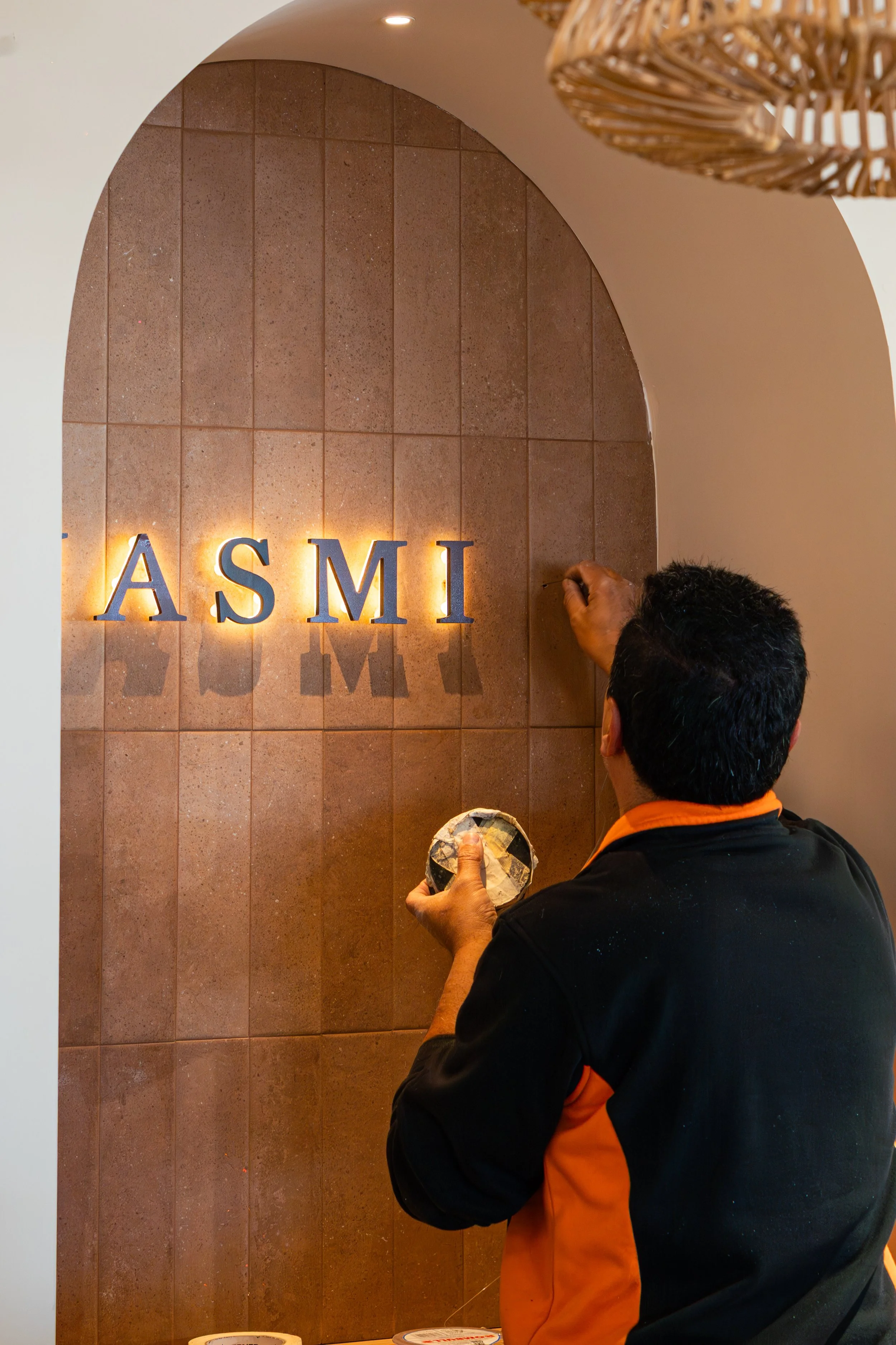 Un trabajador colocando letras iluminadas en la pared, formando la palabra 'ASMI', en un ambiente decorado con paredes de azulejos y una lámpara de techo de fibras naturales.