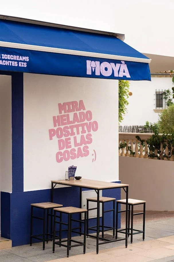 Exterior de una heladería con toldo azul y mesas altas de madera con sillas negras, en la pared hay un cartel rosa con texto en español y un pequeño símbolo de sonrisa.