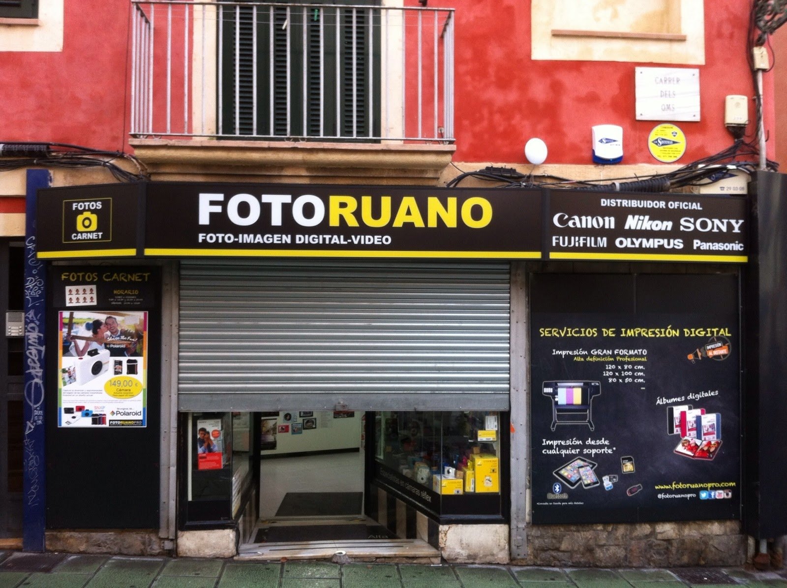 A storefront with a black and yellow sign that reads 'FOTO RUANO', indicating a business specializing in digital photo and video services, as well as digital printing. The store displays logos of cameras and camera brands such as Canon, Nikon, Sony, 
