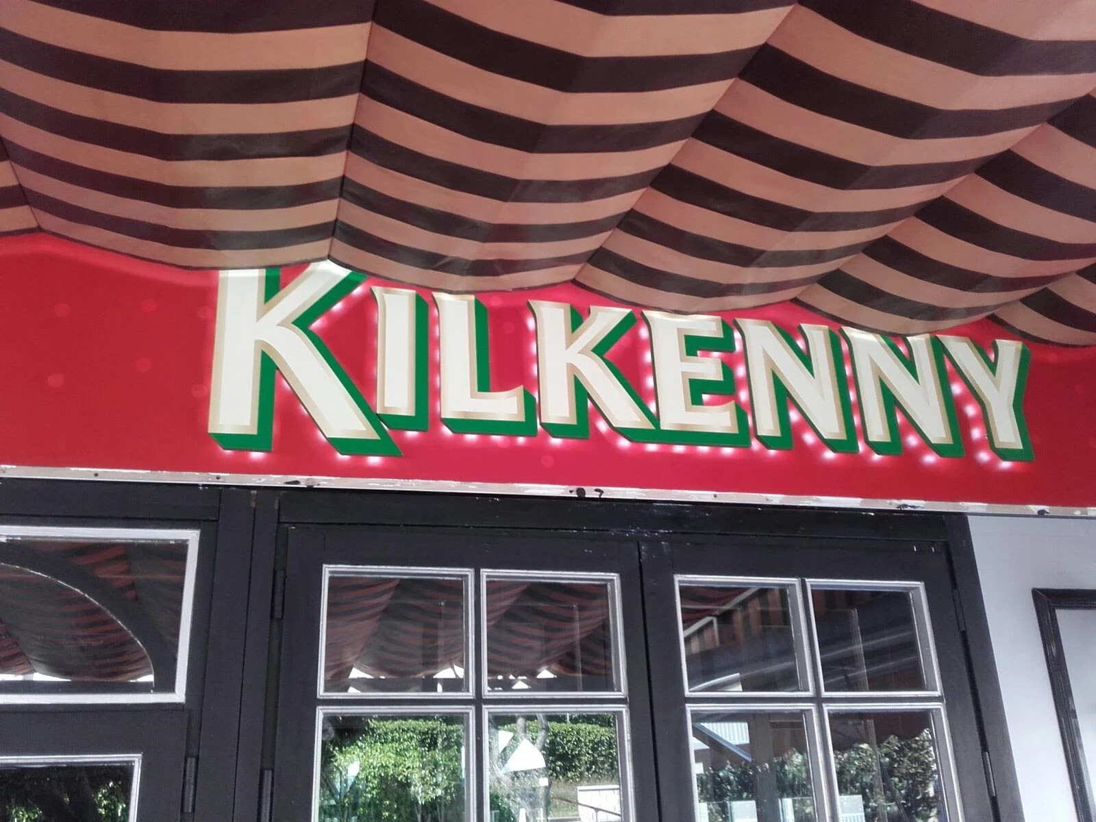 Cartel que dice 'KILKENNY' en letras grandes y blancas con borde verde y sombra roja, con fondo rojo y luces