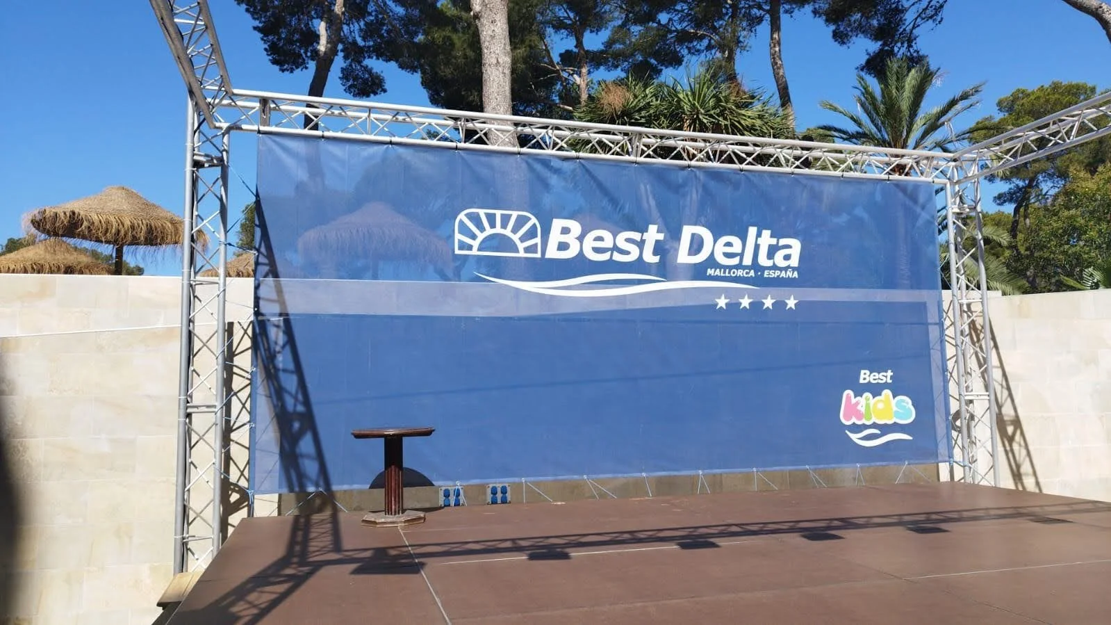 Escenario con cartel de 'Best Delta Mallorca-España' y 'Best Kids', en un espacio al aire libre con sombra de árboles y sombrillas de paja.