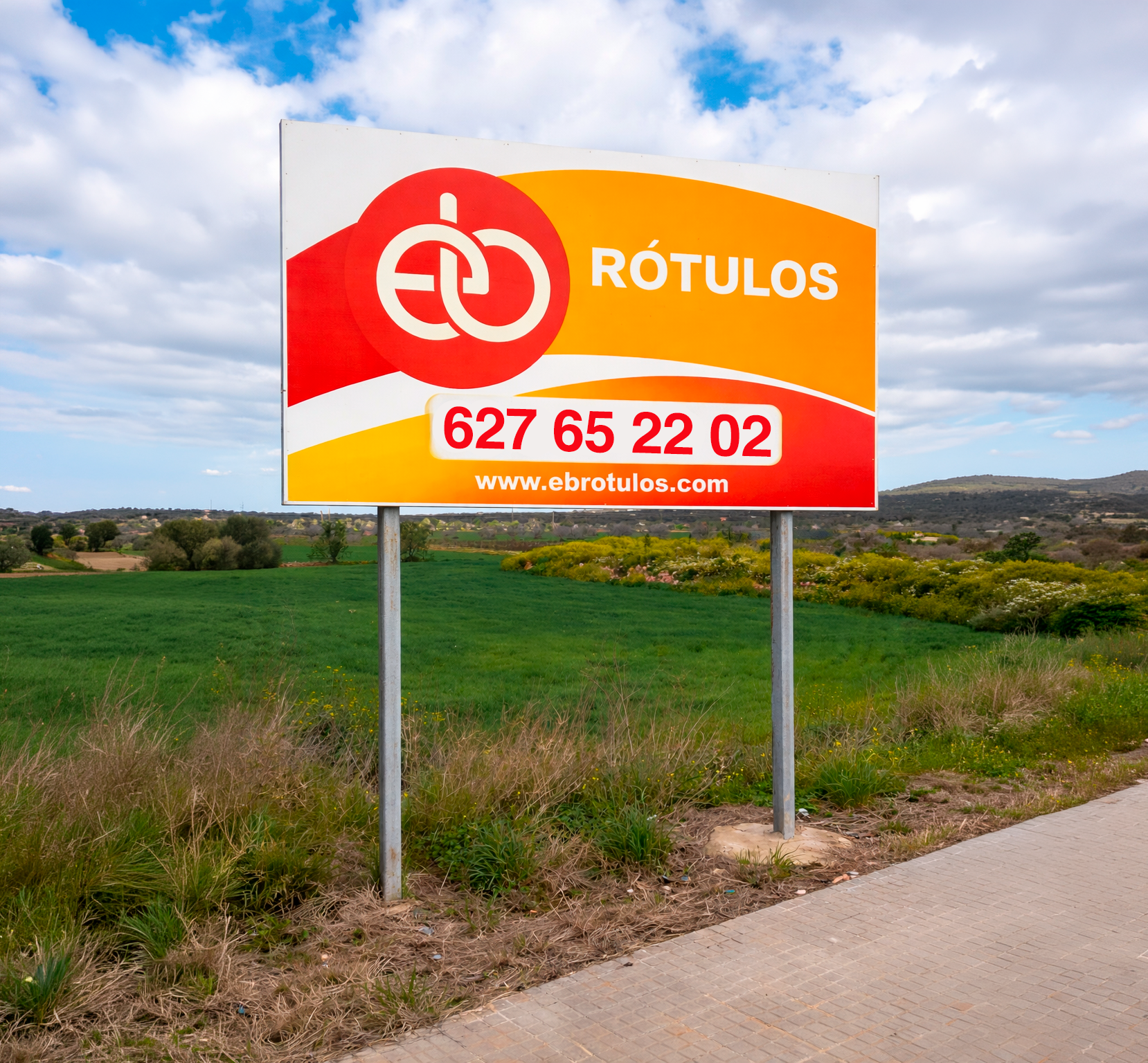 Cartel publicitario de una empresa llamada Rótulos, con un logo de dos anillos entrelazados, y el número de teléfono 627 65 22 02, así como la página web www.ebrotulos.com, en un paisaje rural con cielo nublado, campo verde y colinas a lo lejos.