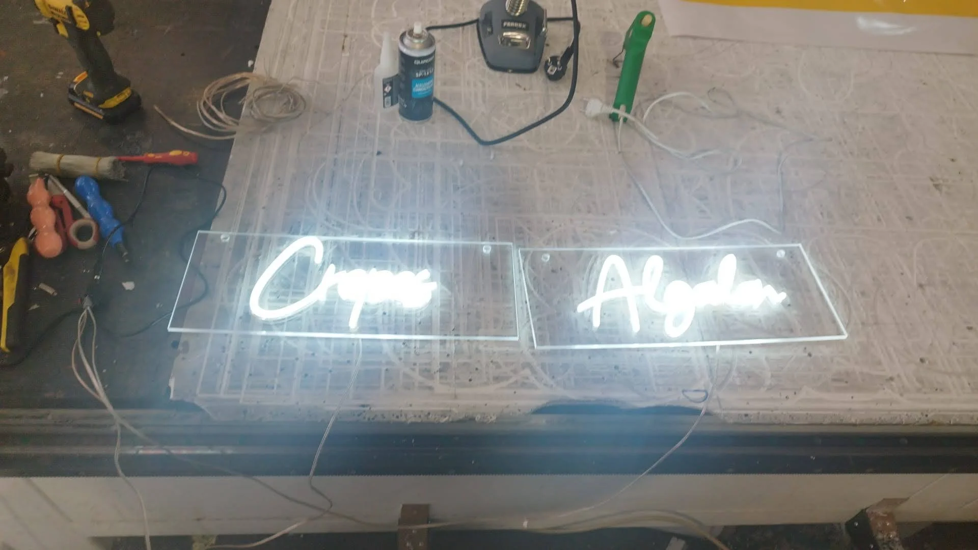 Letrero de neón que dice 'Cerveza' y 'Agua' sobre una mesa de trabajo con herramientas y cables.