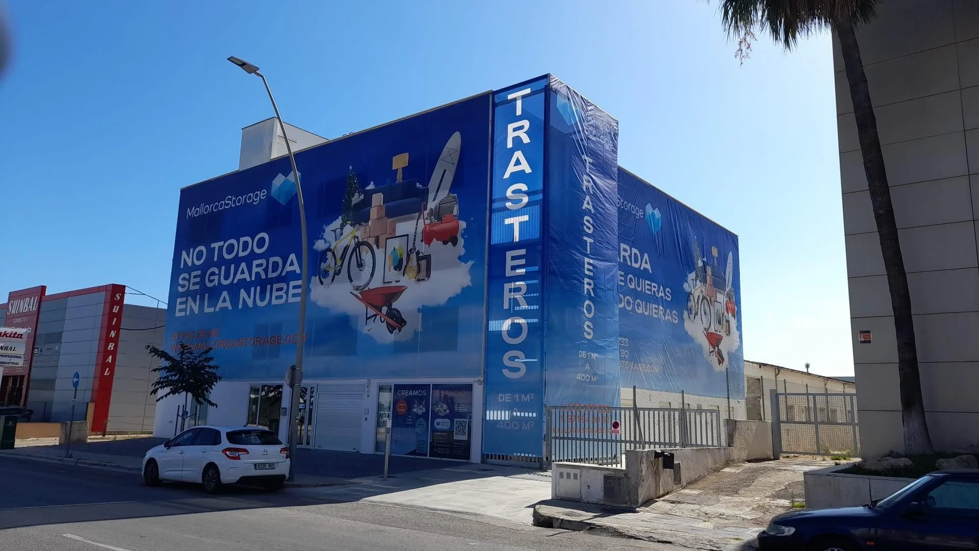 Edificio de almacenamiento con publicidad en español y gráficos de objetos y actividades al aire libre como bicicletas, carretillas y herramientas, con calle, árboles y coches en el entorno.
