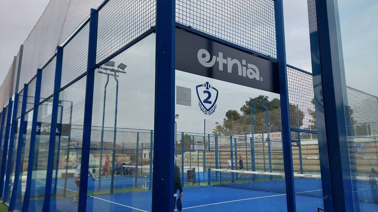 Cancha de pádel techada con paredes de malla metálica azul y piso azul, con jugadores y señales, y un cartel que dice 'etnia'}