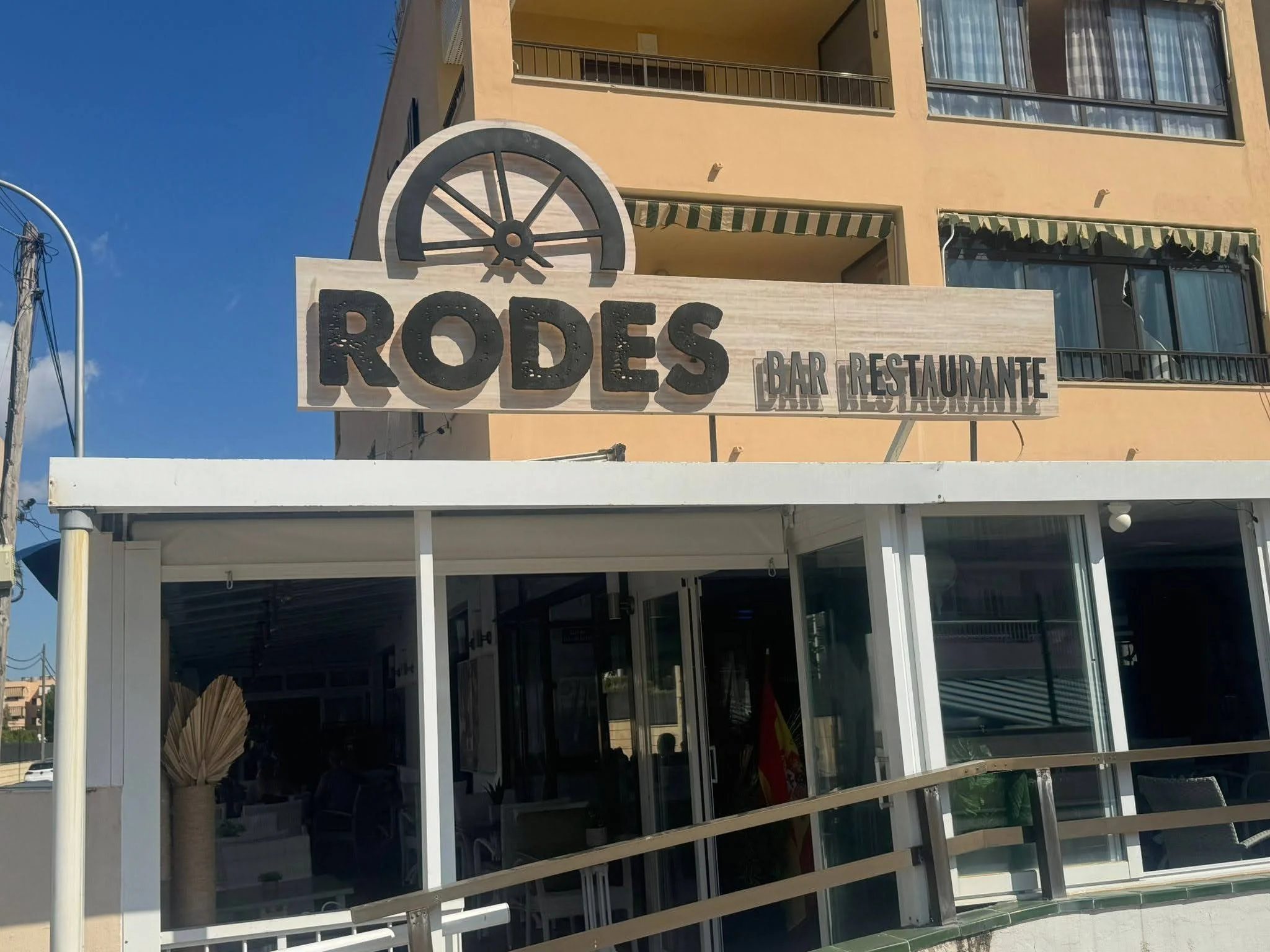 Cartel de un restaurante llamado 'Rodes' con un logotipo de rueda, en un edificio con balcones y ventanas, en un día soleado con cielo azul.