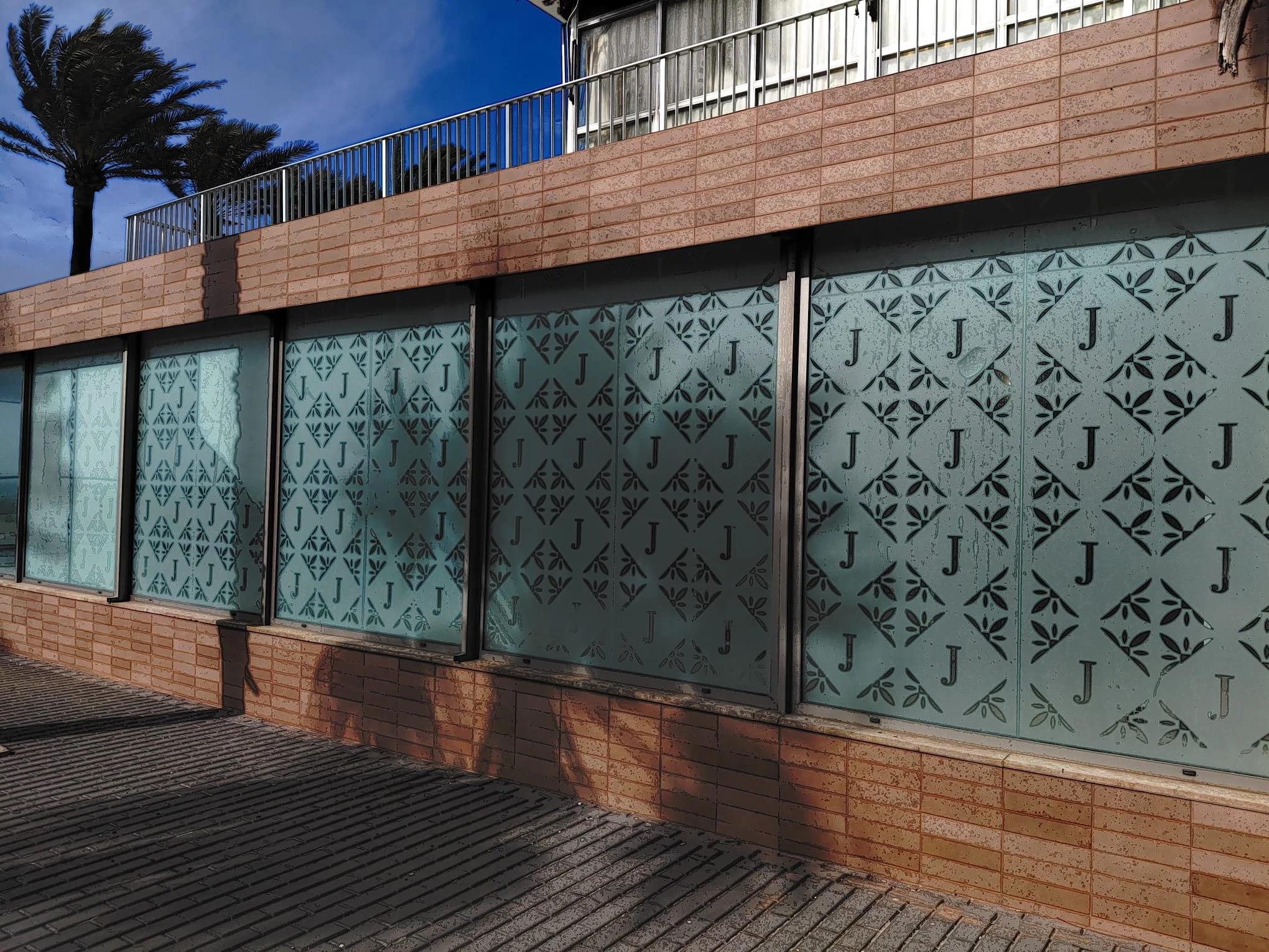Vista de un edificio con ventanas decoradas con patrones de letras J y diseños geométricos, con un muro de ladrillos y sombra en la acera.