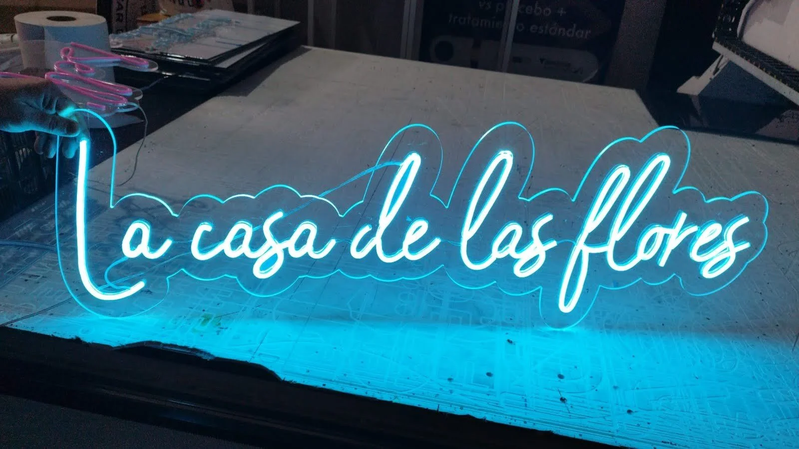 Letrero de neón azul que dice "La casa de las flores" sobre una mesa de trabajo.