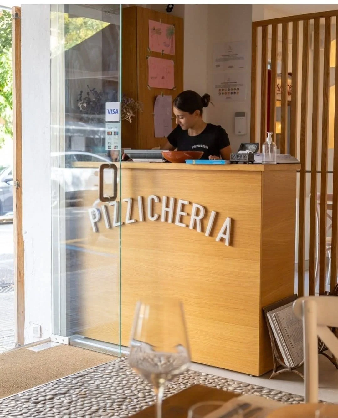 Ventanilla de una pizzería con la palabra 'PIZZERIA' en la parte frontal, vista desde el interior. En el mostrador hay una mujer atendiendo, con una pared de madera detrás y una botella de gel antibacterial en el mostrador.