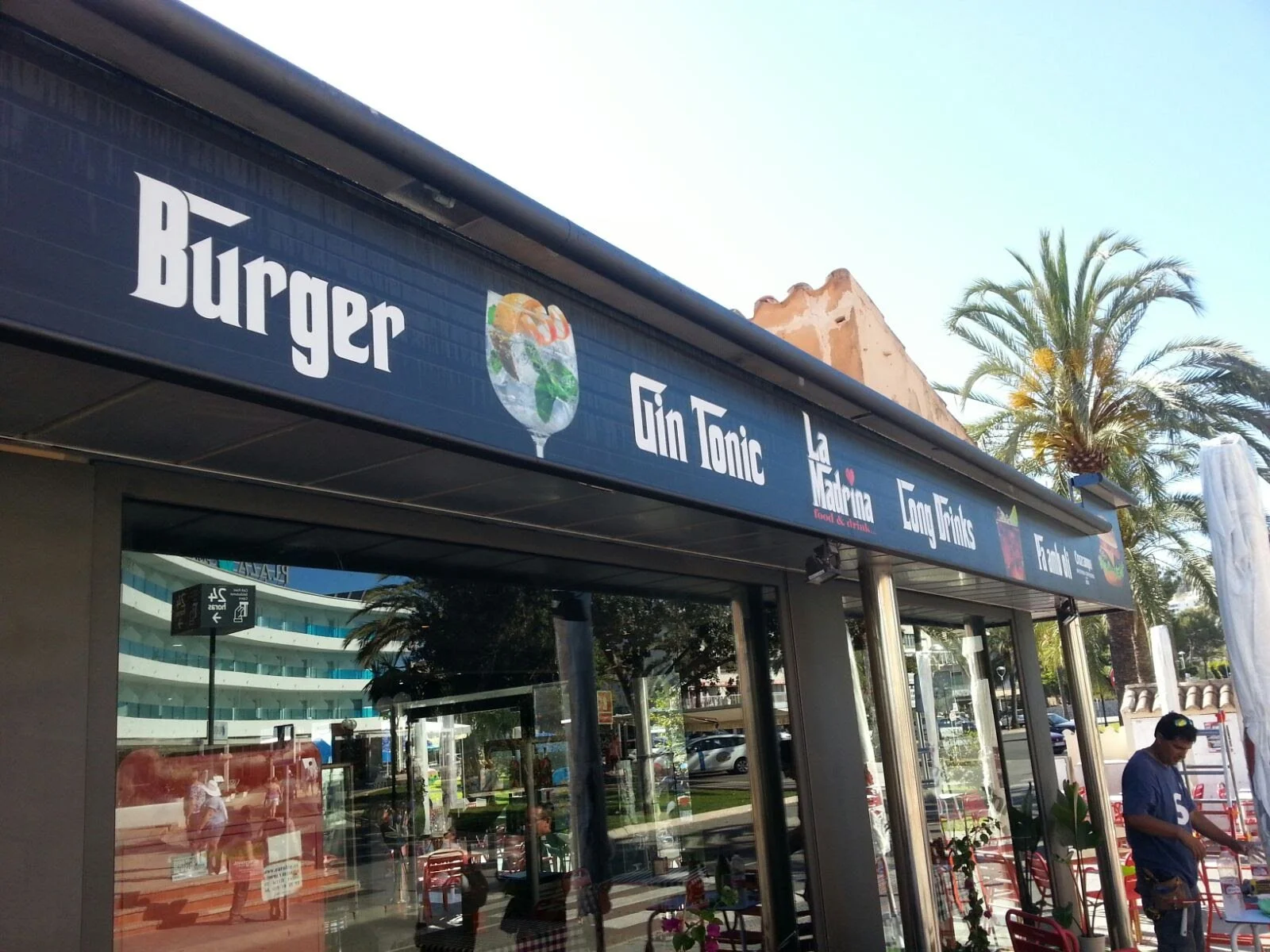 Fachada de una cafetería que ofrece hamburguesas, gin tonic, jugos naturales y cócteles. Se observa parte del establecimiento, algunos clientes y un árbol de palmeras en el exterior.