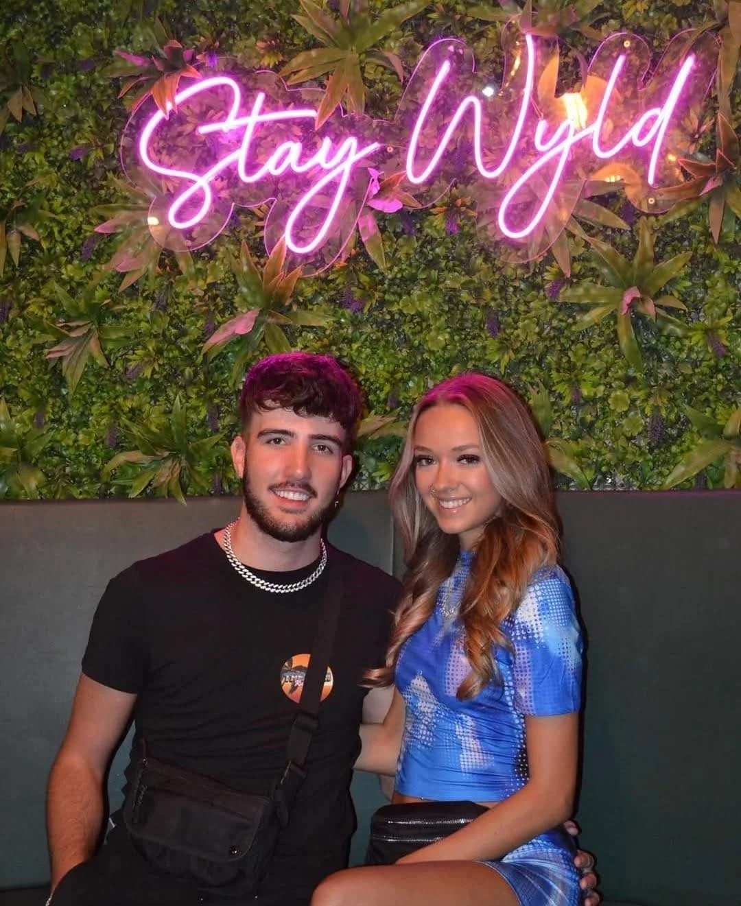 Dos jóvenes hombres y mujeres sentados en un sofá con un fondo de pared cubierta de plantas verdes y una señal de neón rosa que dice "Stay Wyd".