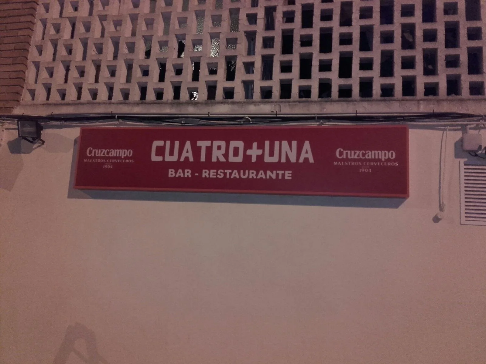Cartel de un bar-restaurante llamado Cuatro + Una, con la marca Cruzcampo, en un muro exterior con ladrillos y bloques de concreto.