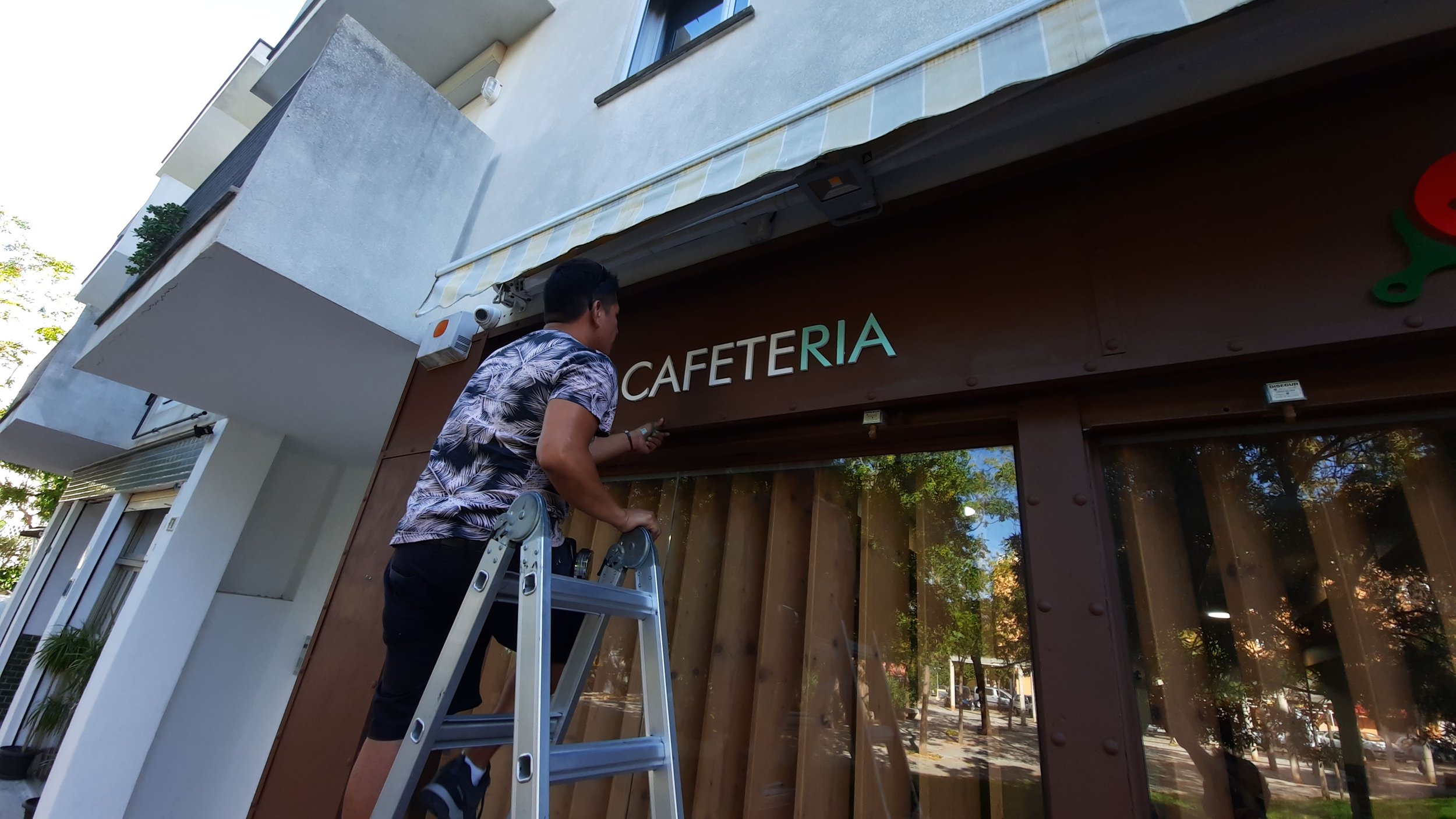 Un hombre colocando una señal que dice 'CAFETERÍA' en la fachada de un establecimiento. Está de pie en una escalera y parece estar haciendo trabajo de mantenimiento o instalación.
