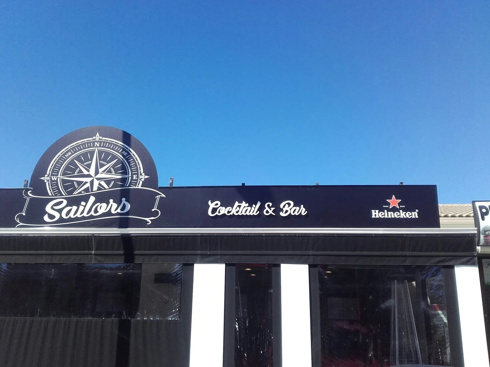 Cartel de un bar y restaurante llamado "Sailors" con un reloj de babor y la frase "Cocktail & Bar" en un fondo negro, y logotipo de Heineken en la esquina derecha