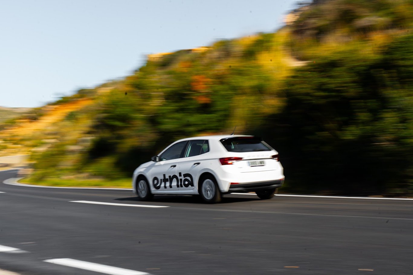 Coche blanco en una curva de carretera, con un logo que dice 'ethnia' en el costado, rodeado de naturaleza con árboles y colinas en el fondo.
