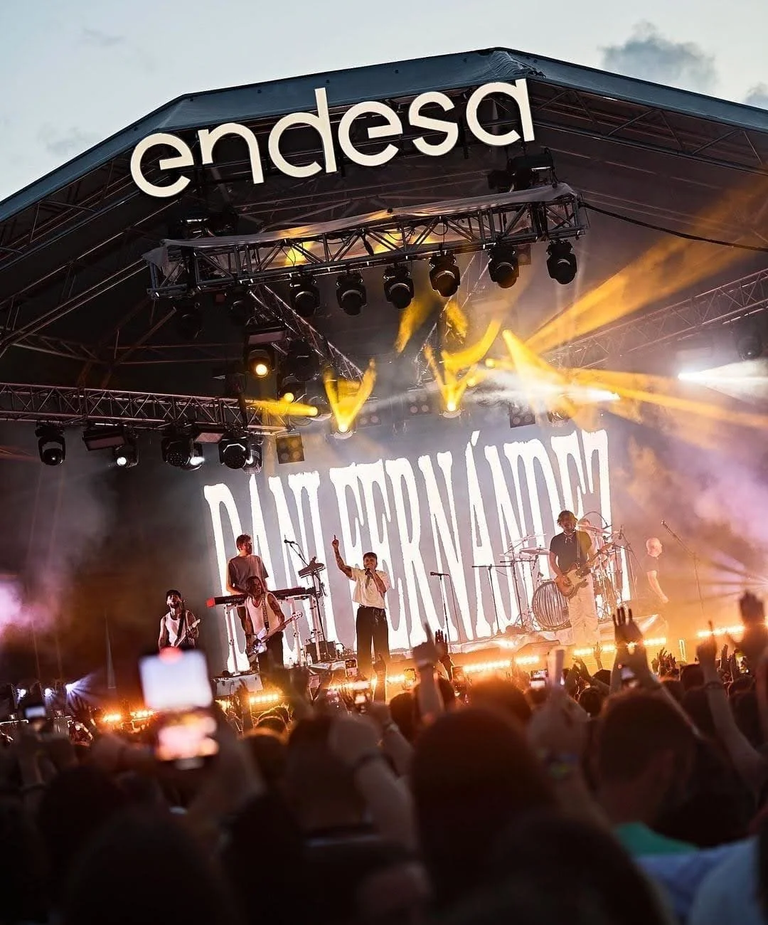 Concierto en un escenario con luces amarillas y banda musical tocando, público en primer plano disfrutando el evento, palabra 'Andesa' en la parte superior del escenario.