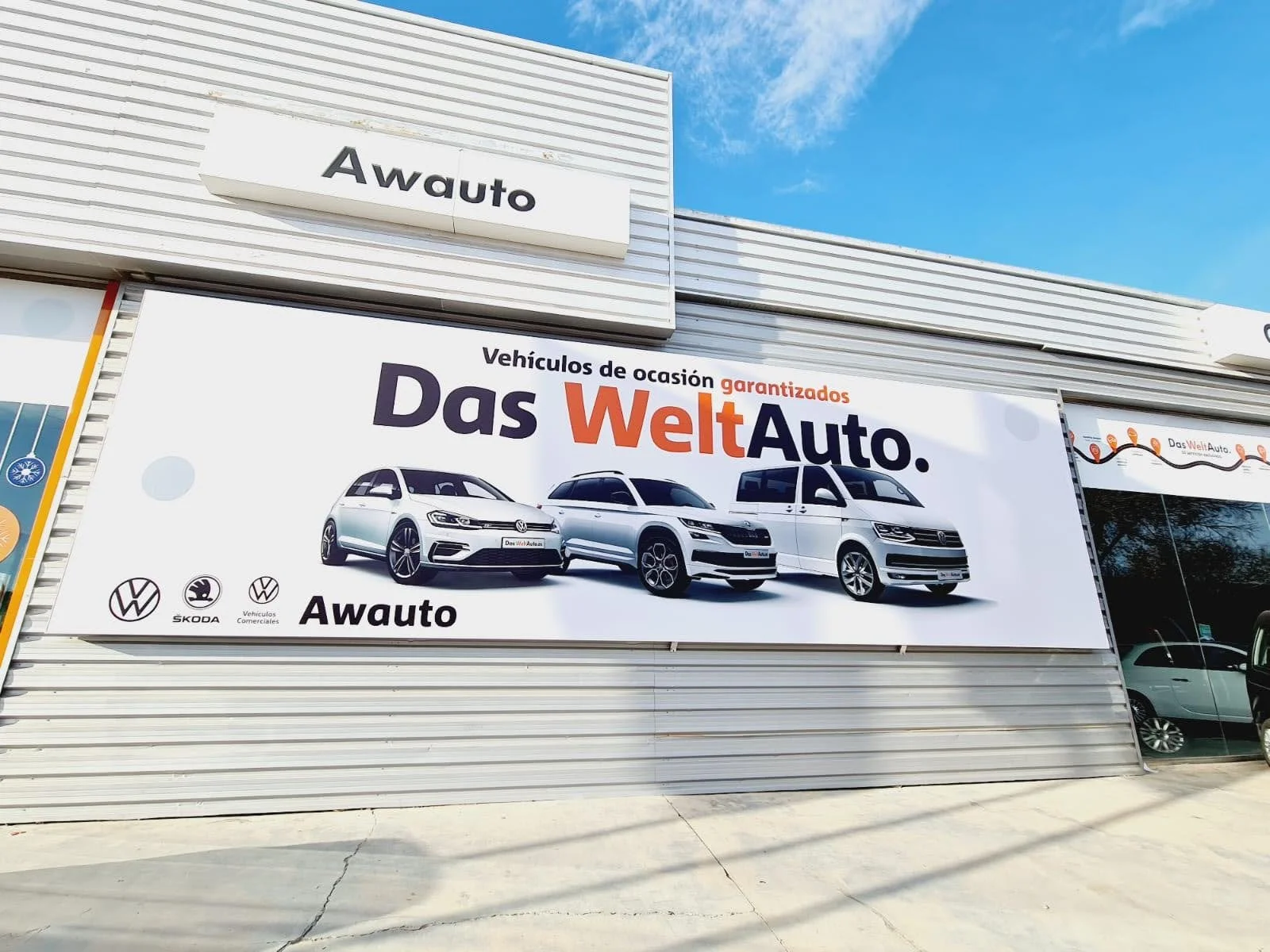 Publicidad en una tienda de autos usados llamada Awauto mostrando tres autos de marca Volkswagen, Skoda y Volkswagen Vehículos Comerciales, con el texto en español 'Vehículos de ocasión garantizados Das WeltAuto.'