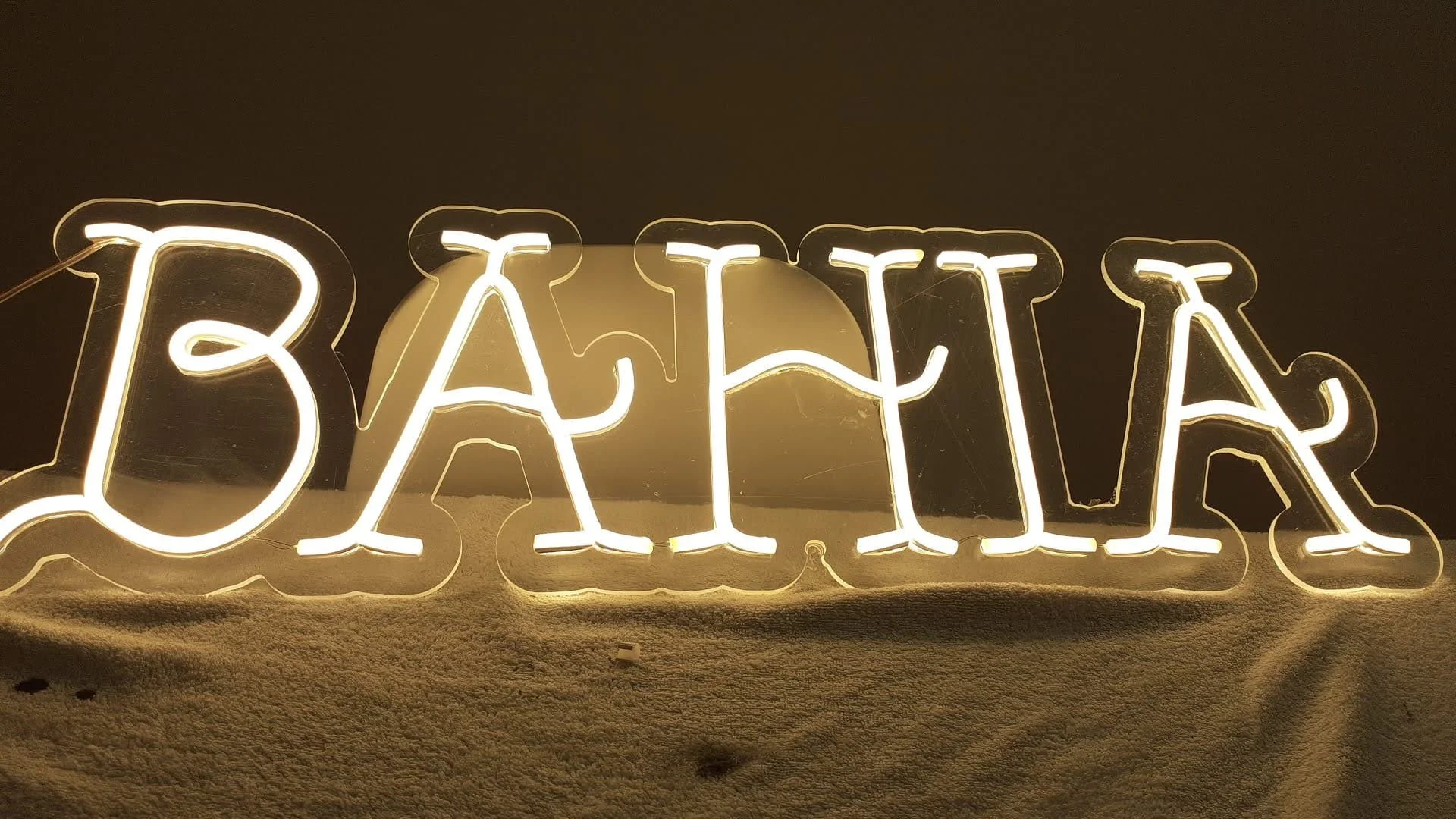 Letrero de neón que dice 'BAR' en letras grandes y blancas, con fondo oscuro y superficie de alfombra beige.