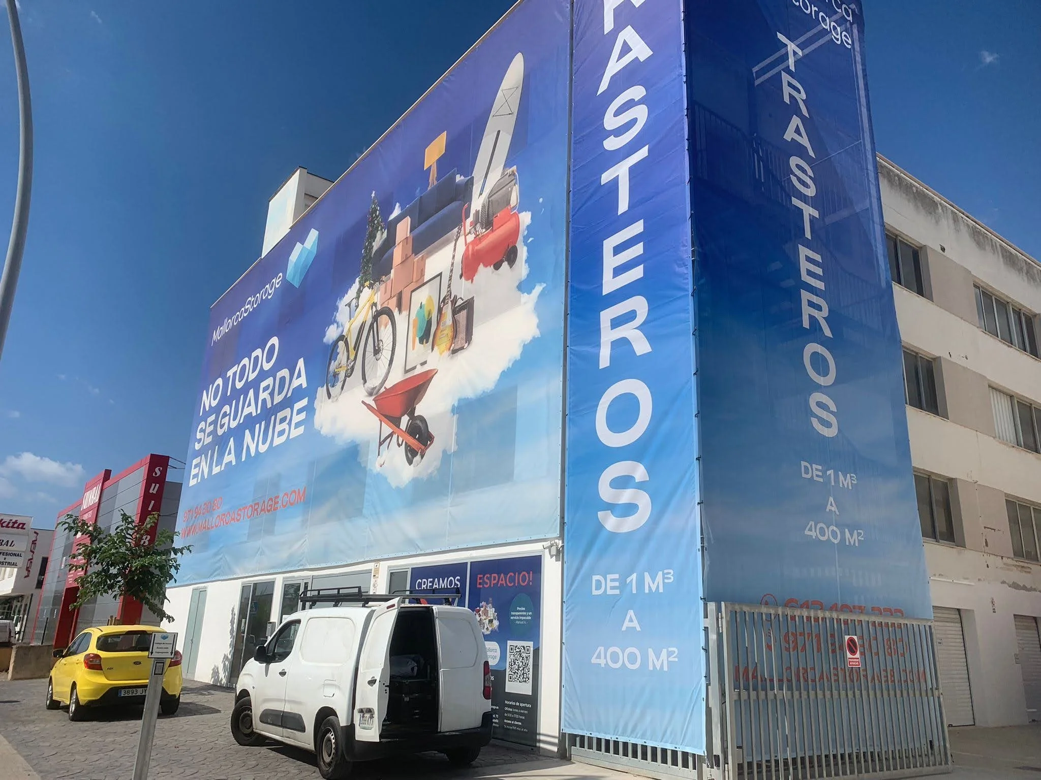 Cartel publicitario azul en un edificio que promociona almacenamiento con la frase "No todo se guarda en la nube" y muestra imágenes de muebles y artículos de mudanza, con detalles en rojo y un código QR, en una calle con varios vehículos y edificios
