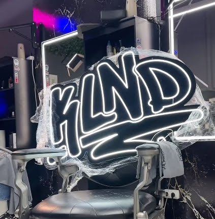 Un letrero de neón que dice 'BLIND', en un establecimiento de barbería o salón de belleza, con un espejo y silla en primer plano.