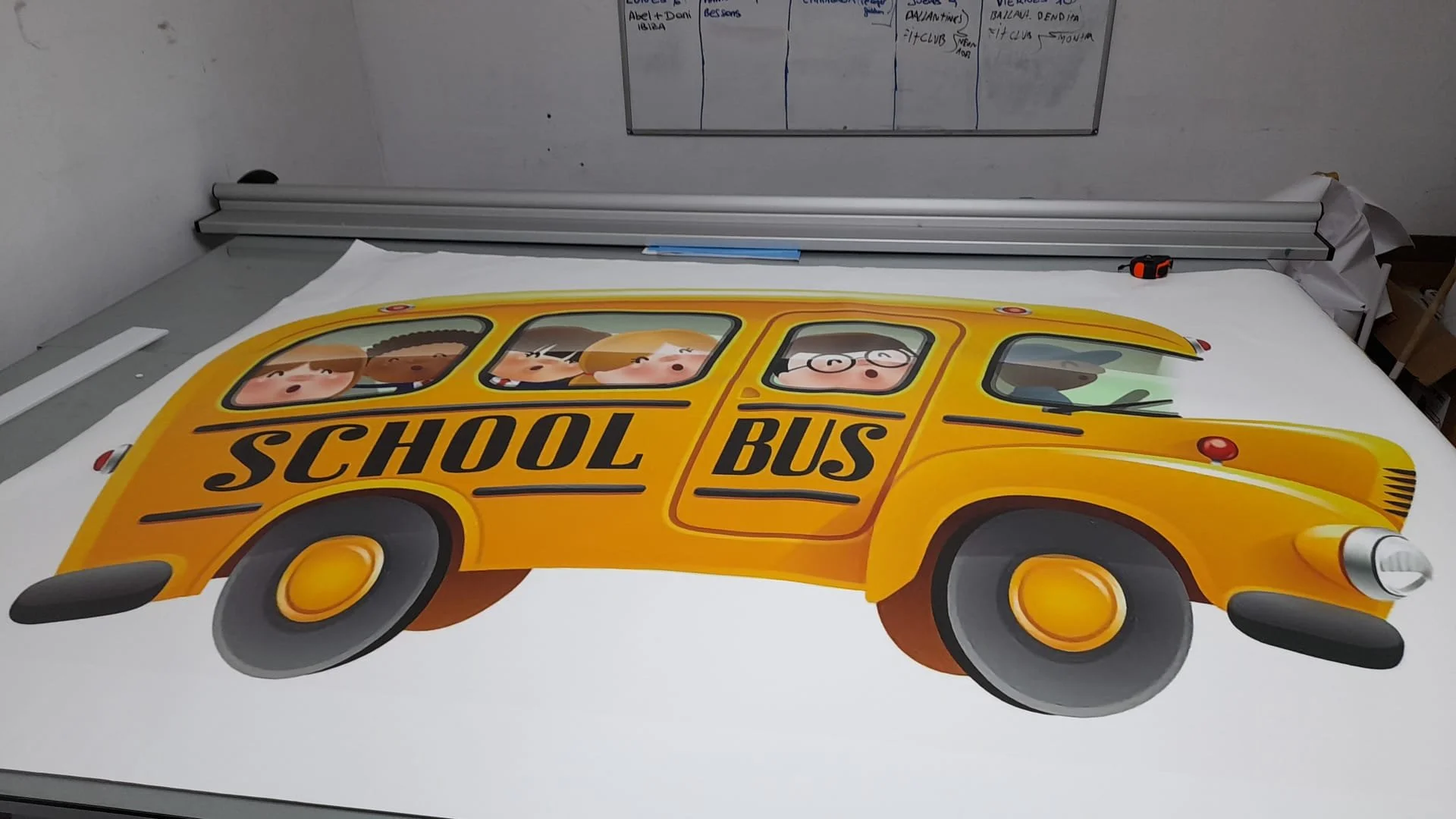 Dibujo de un autobús escolar amarillo con niños en las ventanas, en un taller con una mesa, reglas y una pizarra en el fondo.