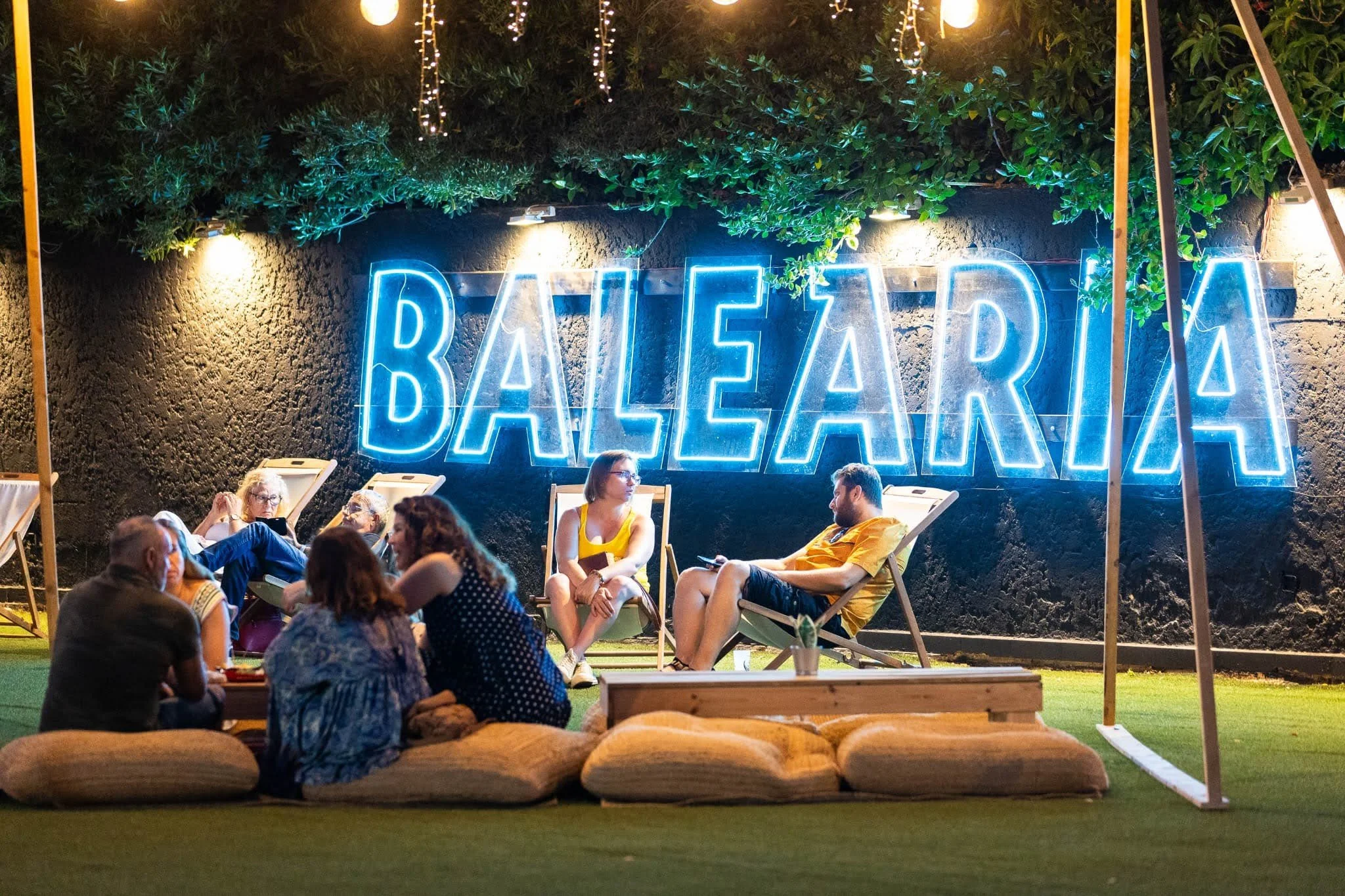 Un grupo de personas sentado en sillas y bancos en un espacio de exterior iluminado con luces, con un cartel de neón azul que dice 'Balearia'.