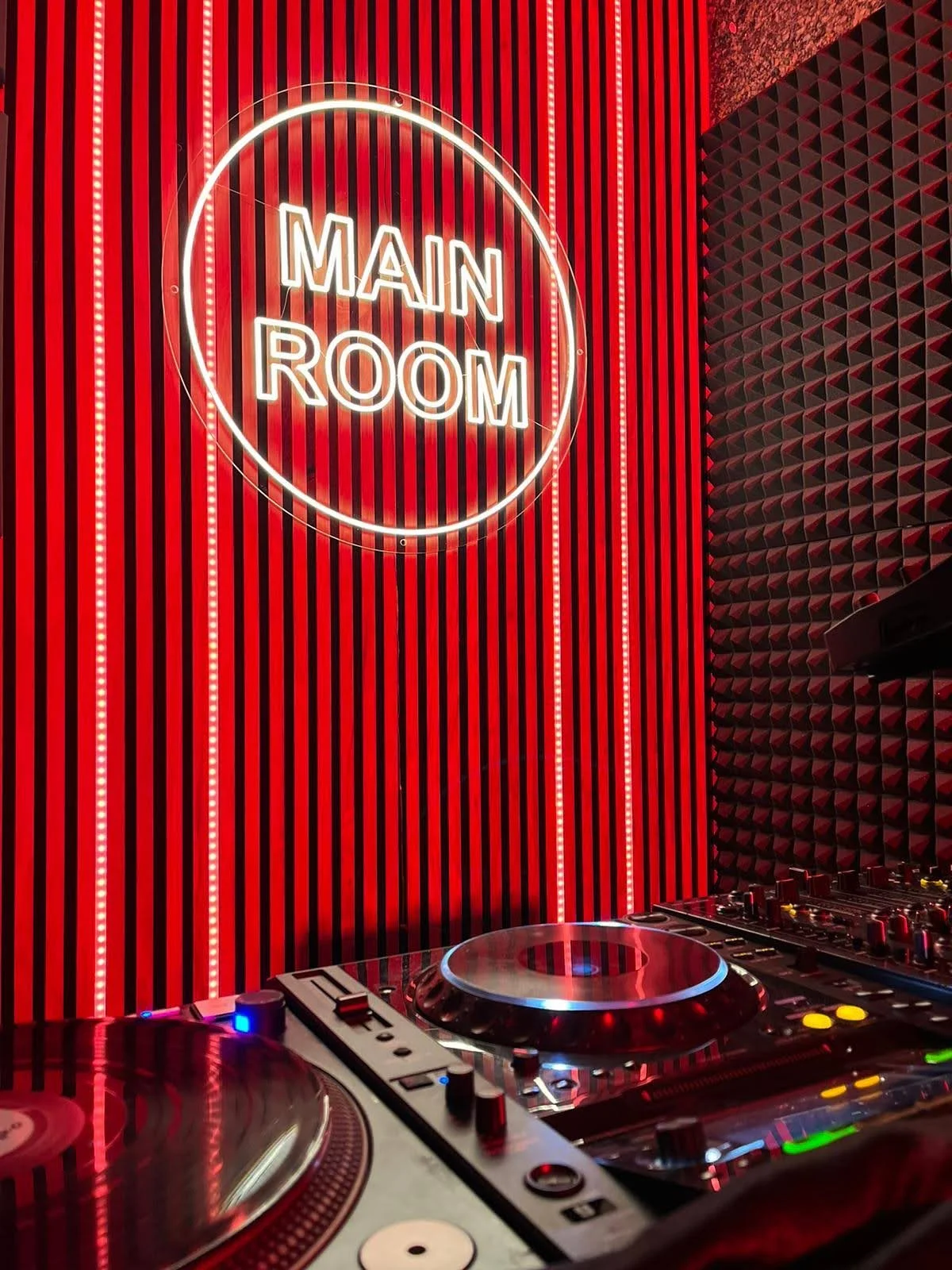 Cartel de neón que dice 'Main Room' en un fondo de paredes rojas con rayas verticales y paneles de espuma acústica negra.