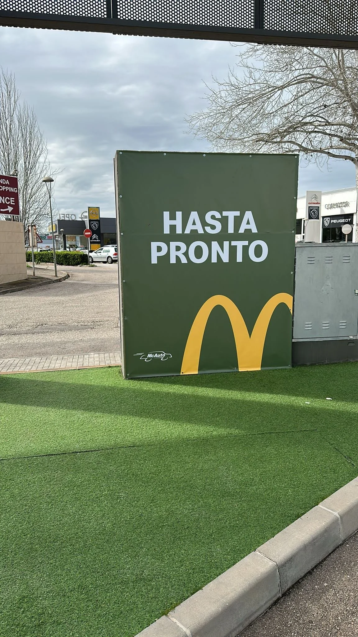 Cartel de McDonald's que dice 'Hasta Pronto' en color verde con el logo de McDonald's en la parte inferior y un fondo de estacionamiento con árboles y edificios.