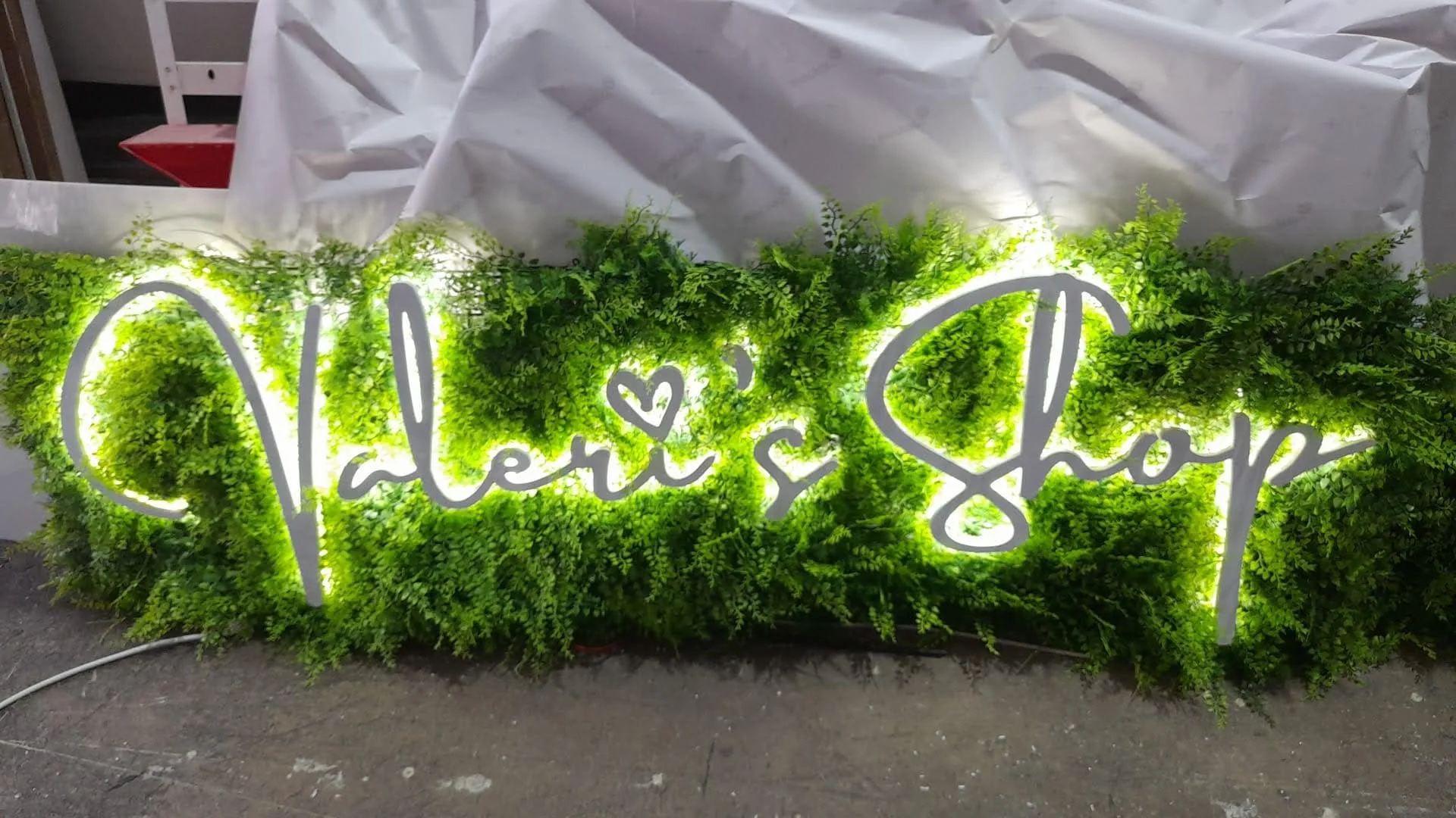 Letrero luminoso que dice 'Valenti Shop' rodeado de plantas verdes.