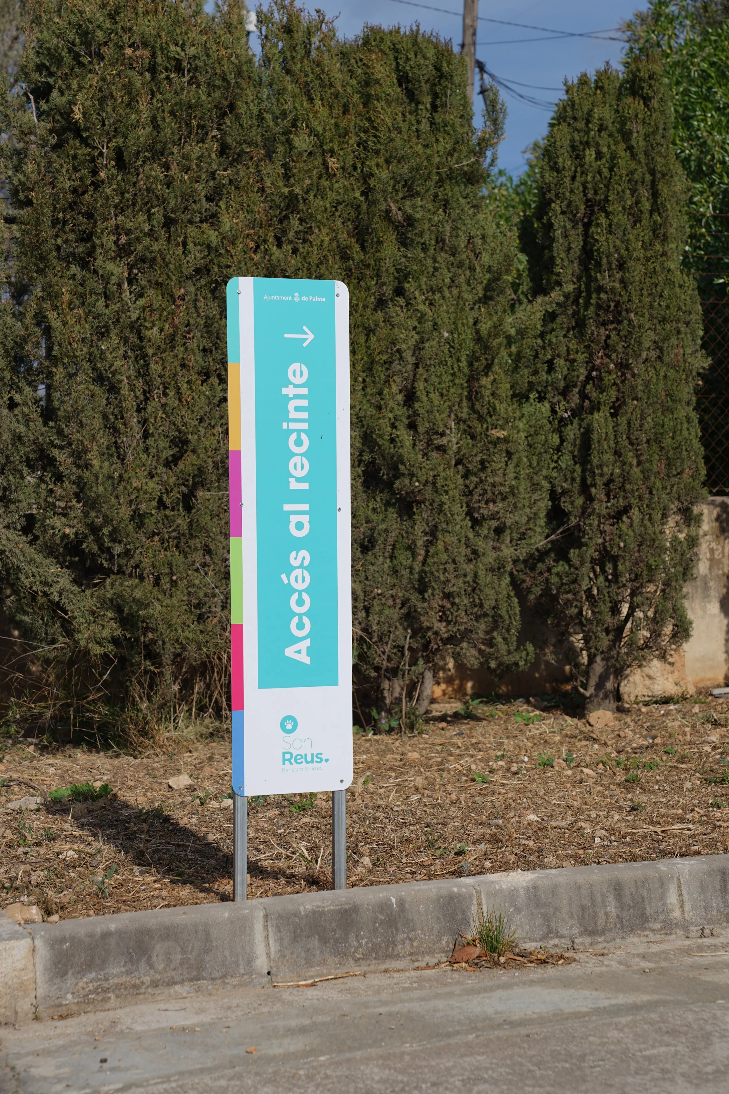 Cartel de acceso con flecha, en un entorno con arbustos y árboles. La señal dice 'Accés al recinte' con una flecha hacia la derecha.
