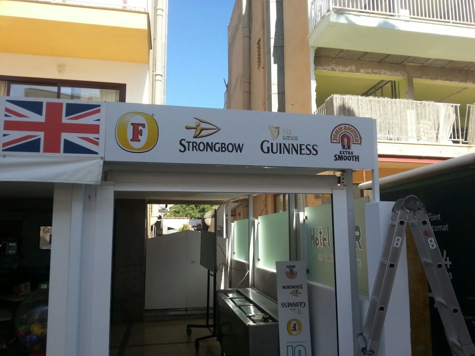 Estructura de una especie de cabina o quiosco con publicidad de marcas de cerveza y a la izquierda una bandera del Reino Unido.