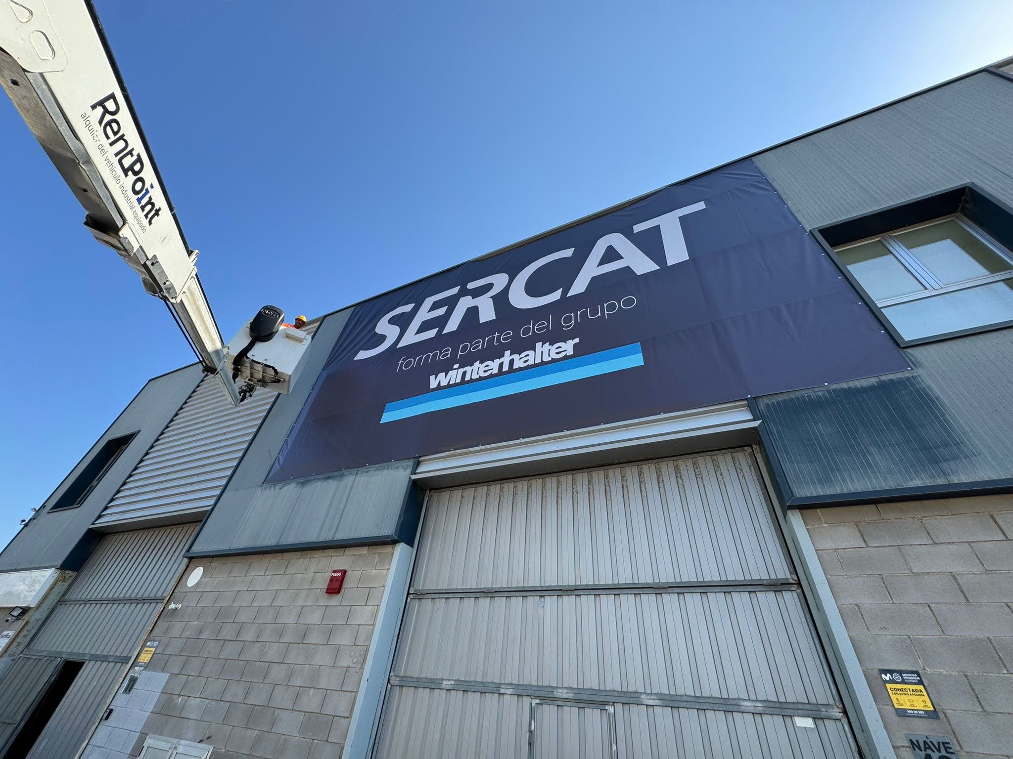 Vista de un edificio de negocios con un cartel grande que dice 'SERCAT' y la frase 'forma parte del grupo Winterhalter', además de una grúa en la parte izquierda de la imagen.