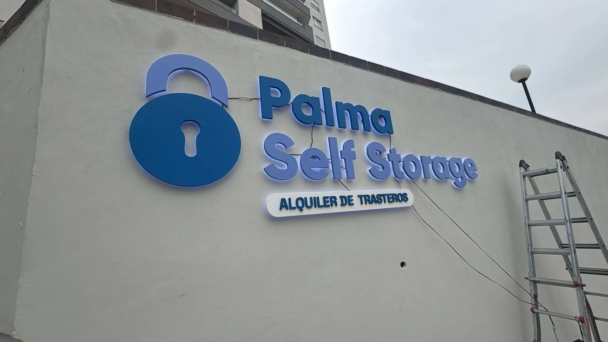 Se observa un letrero iluminado en una pared que dice 'Palma Self Storage' y debajo en letras más pequeñas 'Alquiler de Trasteros', con un símbolo de candado en el lado izquierdo y una escalera a la derecha.