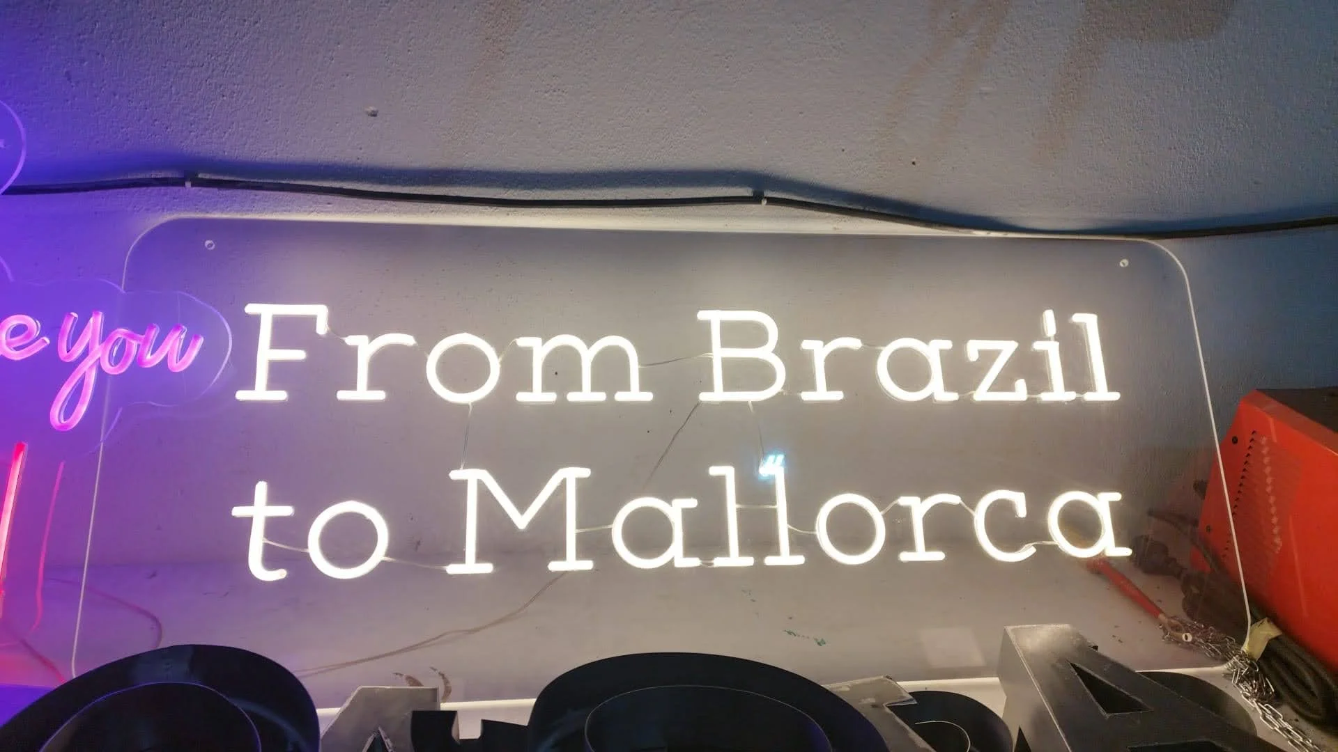 Letrero de neón que dice 'from Brazil to Mallorca' en letras blancas, con parte del fondo en tonos morados y una parte de la decoración en la esquina inferior derecha de color naranja.