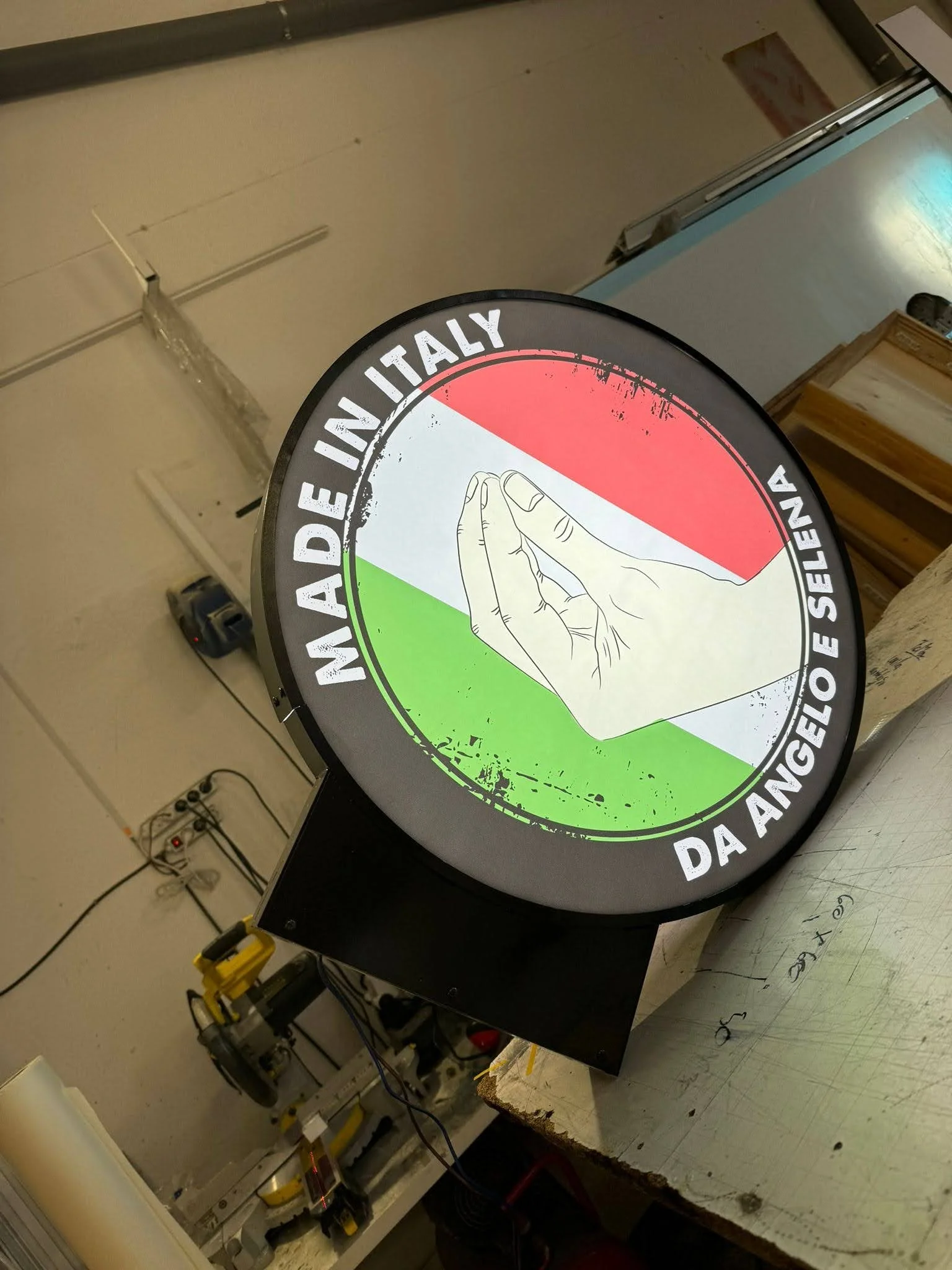 Cartel circular con los colores de la bandera de Italia, con la imagen de una mano realizando un gesto de oración y el texto en italiano y español para promover la adopción de mascotas. Incluye la frase en italiano 'Adottare in Italia' y en español '