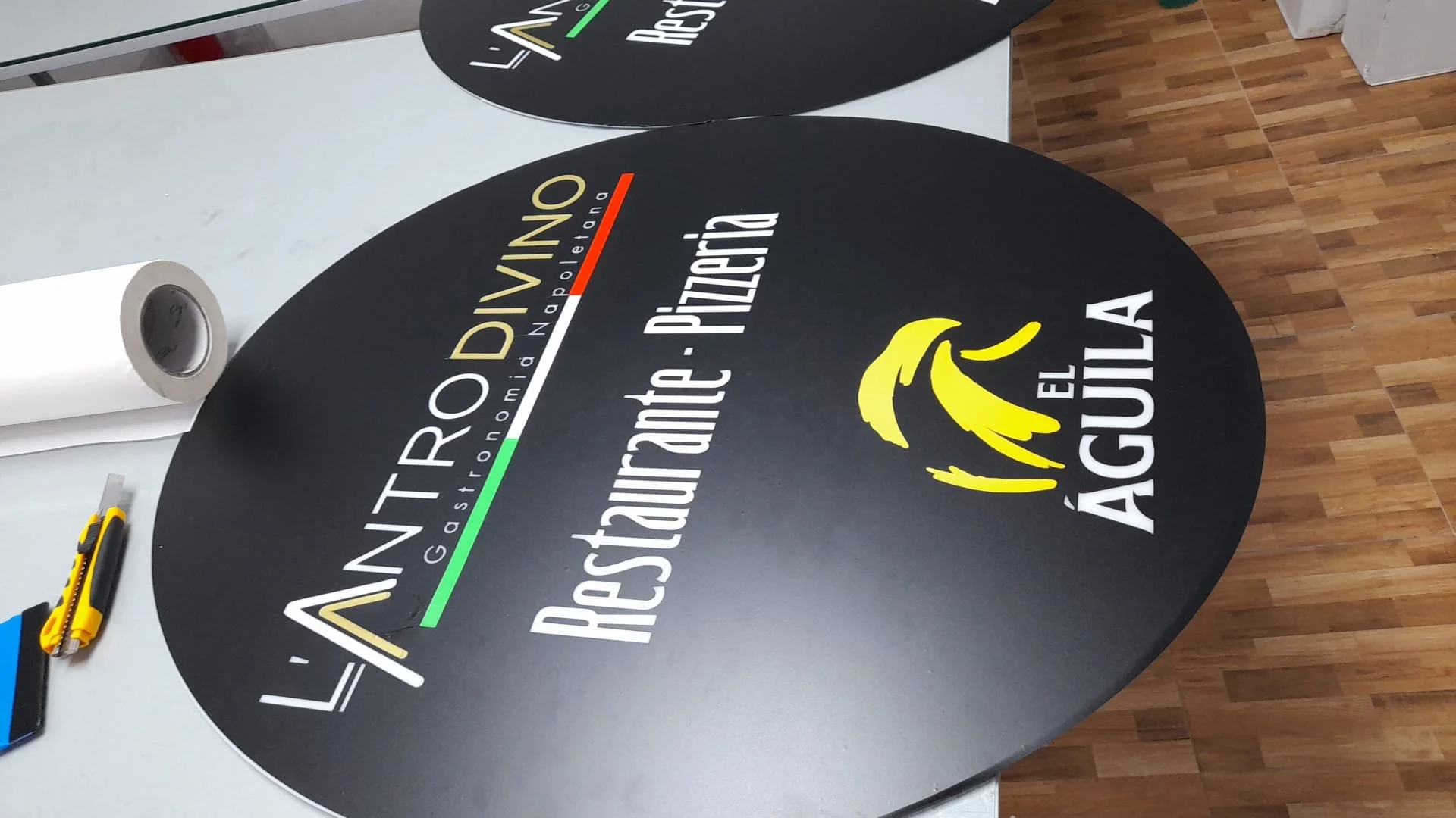 Cartel ovalado negro con logotipo y texto en colores blanco, amarillo, rojo y verde en una mesa, en un entorno de oficina o comercio.