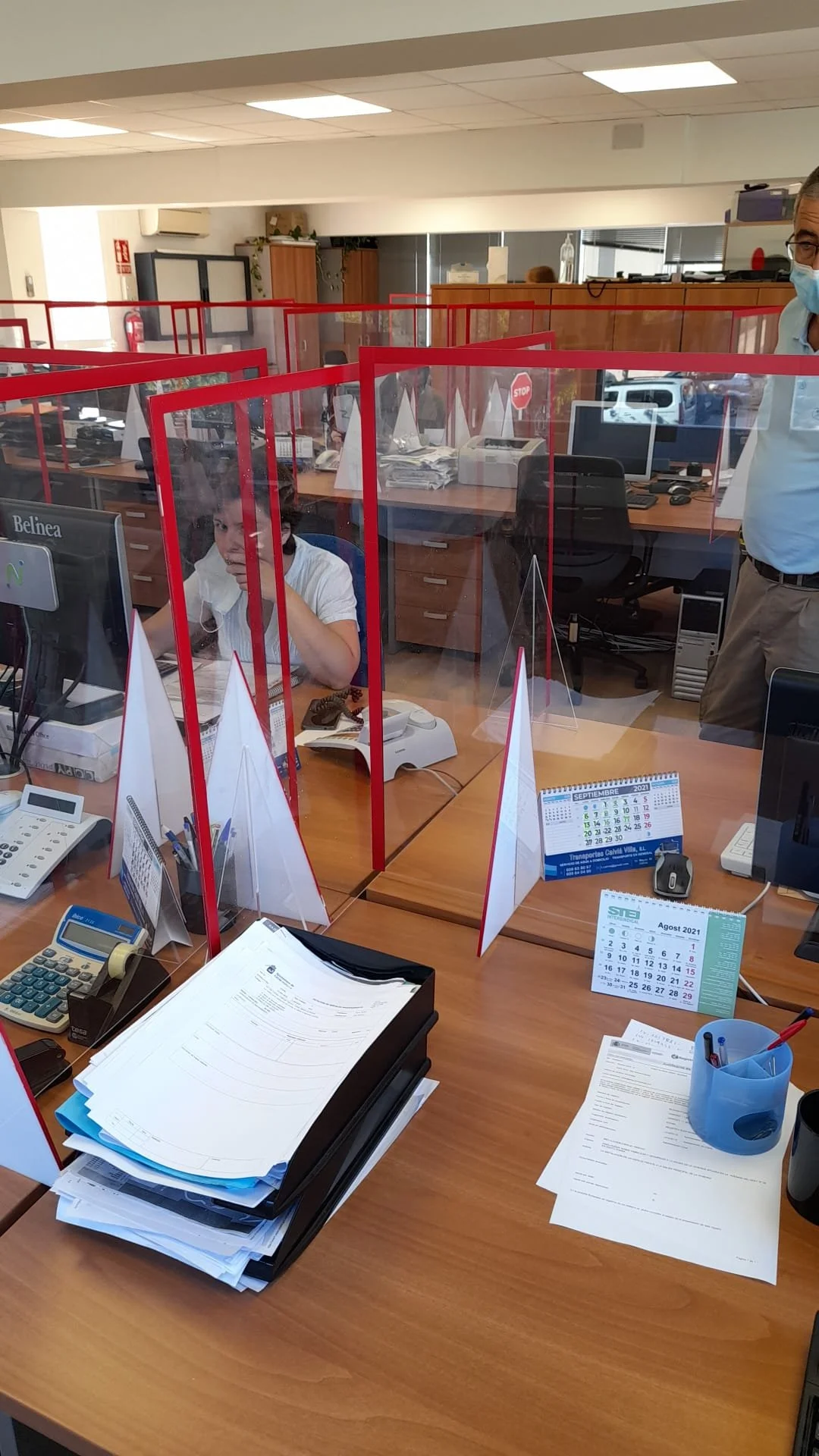 Oficina con escritorios y escritorios con pantallas y algunos documentos, con barreras de acrílico para protección. Se ven dos empleados, uno en el fondo y uno en primer plano, usando mascarillas.