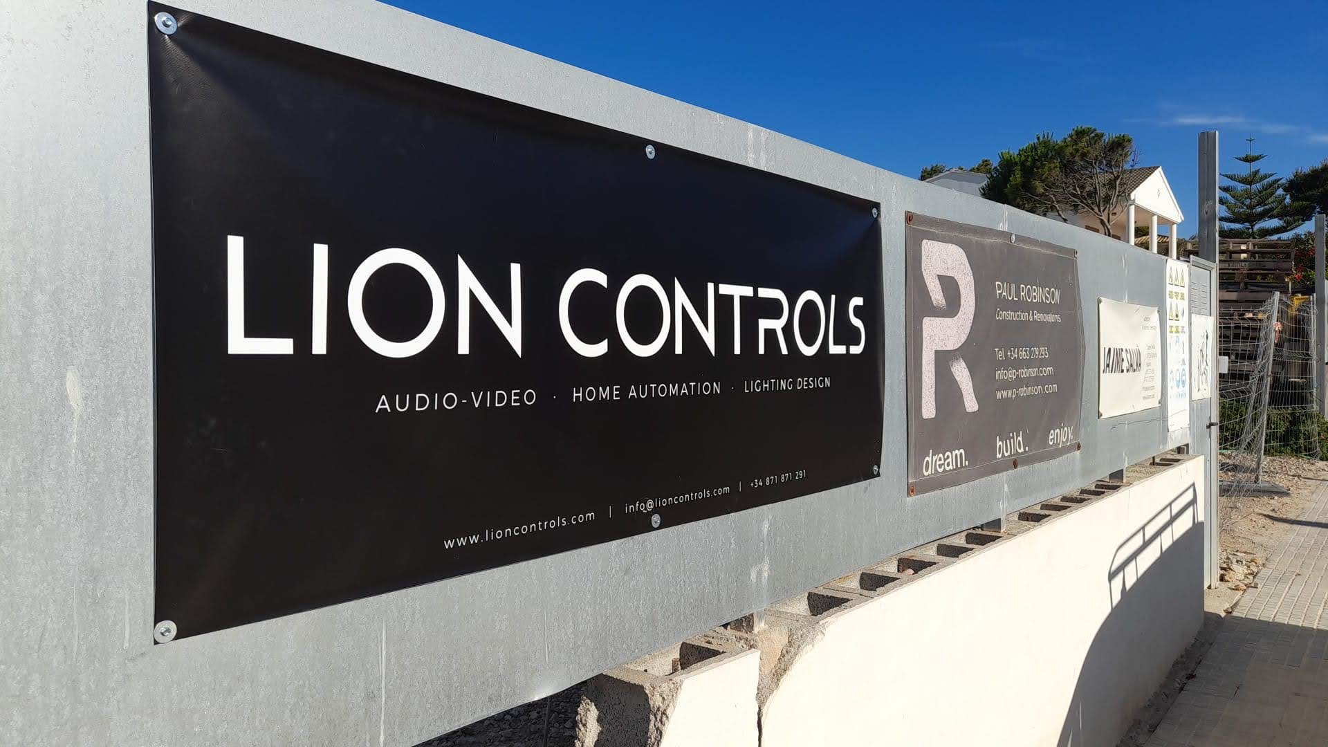 Carteles publicitarios en una pared de concreto: uno que dice 'Lion Controls' con información sobre audio, video, domótica y diseño de iluminación; otro con datos de contacto y slogans como 'dream', 'build', 'enjoy'.