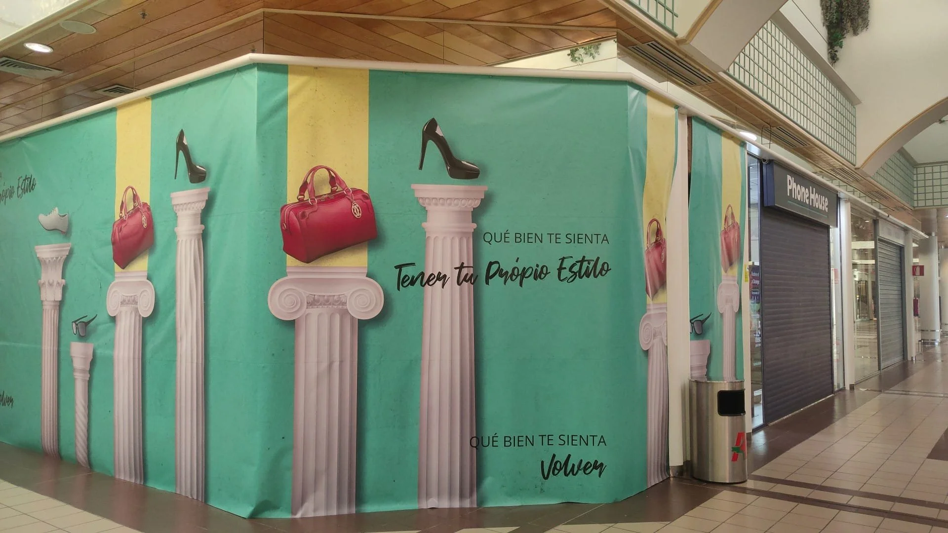 Cartel en centro comercial con columnas griegas y bolsas de mano, con texto en español que dice 'Qué bien te sienta tener tu propio estilo' y elementos decorativos en el fondo.