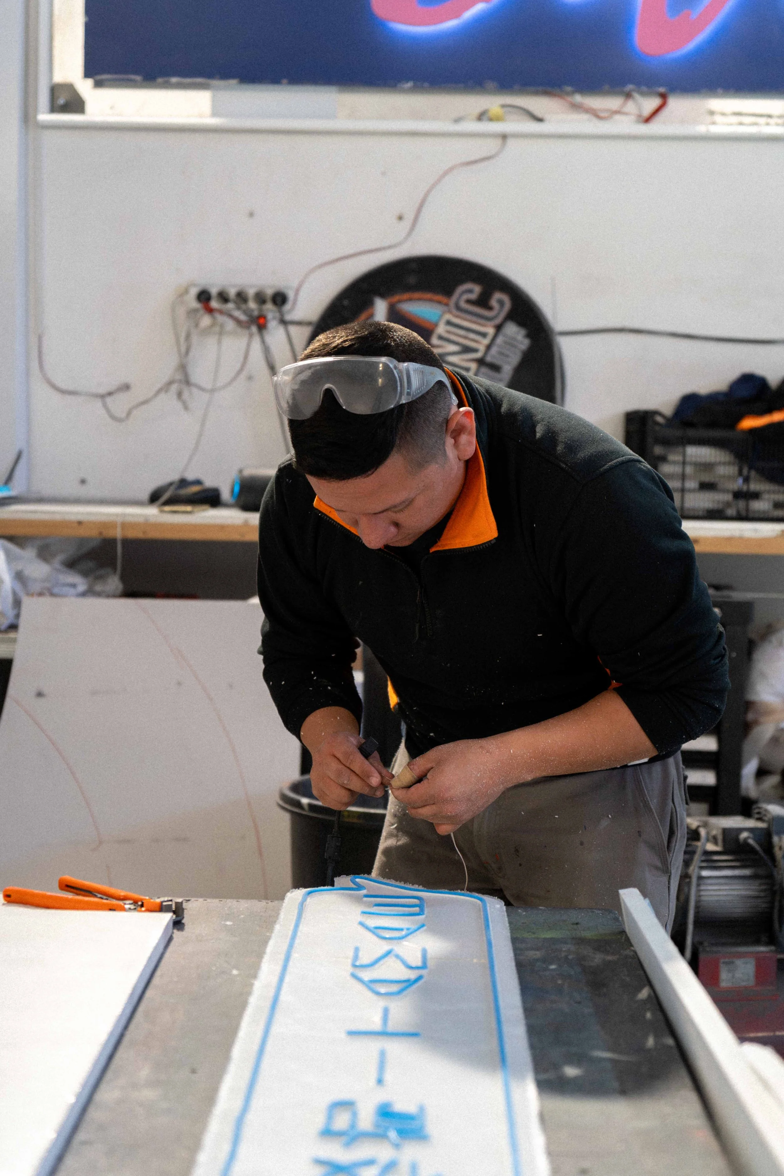 Un hombre trabajando en una mesa, usando gafas protectoras, haciendo detalles con un bolígrafo sobre una pieza de arte o señal artesanal en un taller.