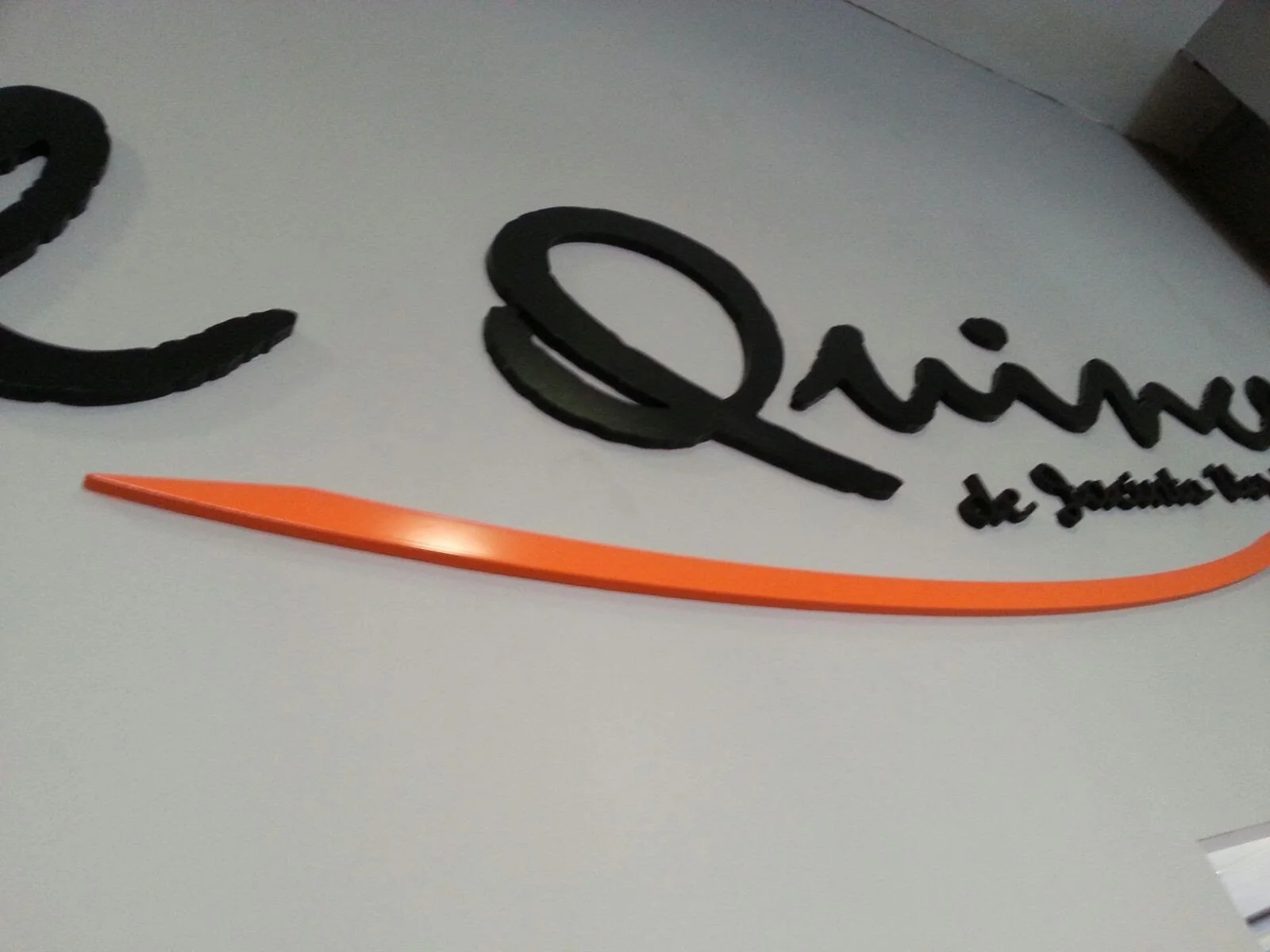 Se ve una pared blanca con letras negras en relieve que forman la palabra 'Qimis' y un texto más pequeño debajo que dice 'de Sporttin'. Además, hay una línea curva naranja debajo del texto.