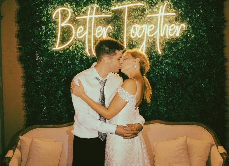 Pareja joven besándose en ceremonia con fondo de muro de plantas y un letrero de neón que dice 'Better Together', en un espacio decorado con sillón y cojines.