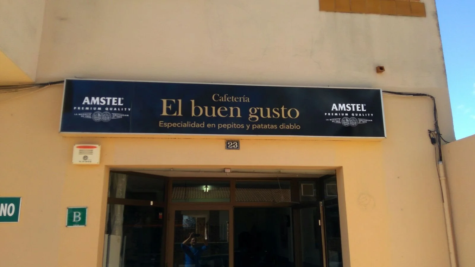 Cartel de un café llamado 'El buen gusto' que especifica su especialidad en pepitos y papas diablos, en un edificio con paredes beige y una puerta de vidrio.