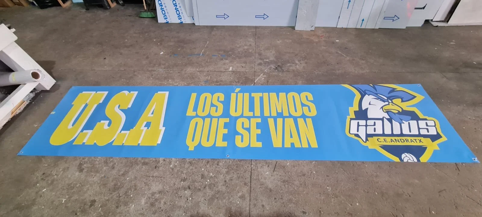 Cartel azul con letras amarillas y blancas que dice: "U.S.A Los últimos que se van" y muestra el escudo del equipo de fútbol C.E. Andratx con un águila estilizada en colores azul, blanco y amarillo.