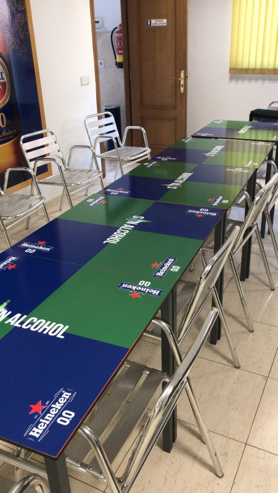 Mesa de reunión con un tapete de cartón con marca de cerveza Heineken. Sillas metálicas alrededor de la mesa en una sala interior con puerta de madera, bandera amarilla y cartel en la pared.
