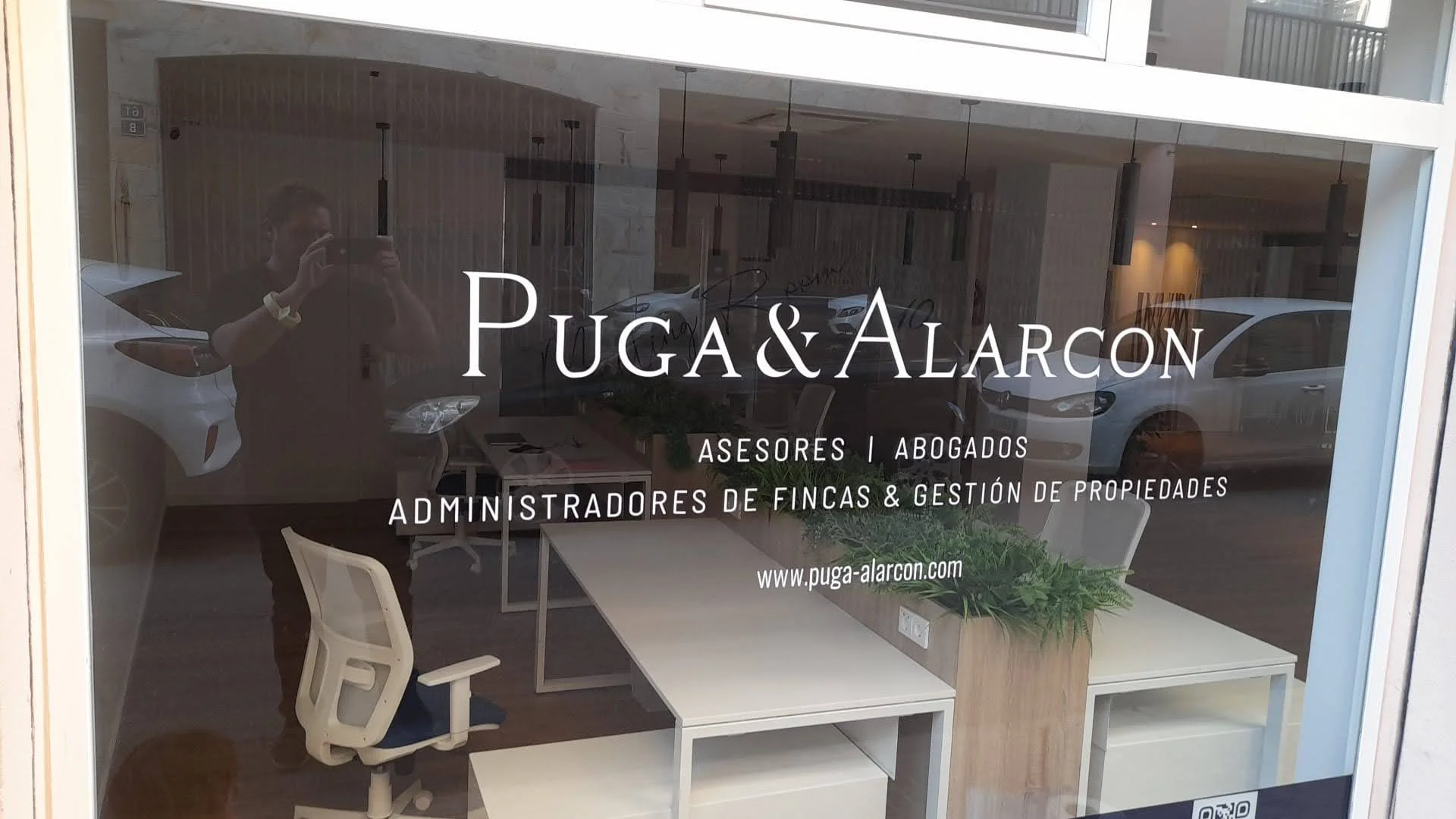 Ventana de oficina que muestra el logotipo de Puga & Alarcon, una firma de abogados y asesores en bienes raíces y gestión de propiedades, con una sala de reuniones y autos visibles en el reflejo.