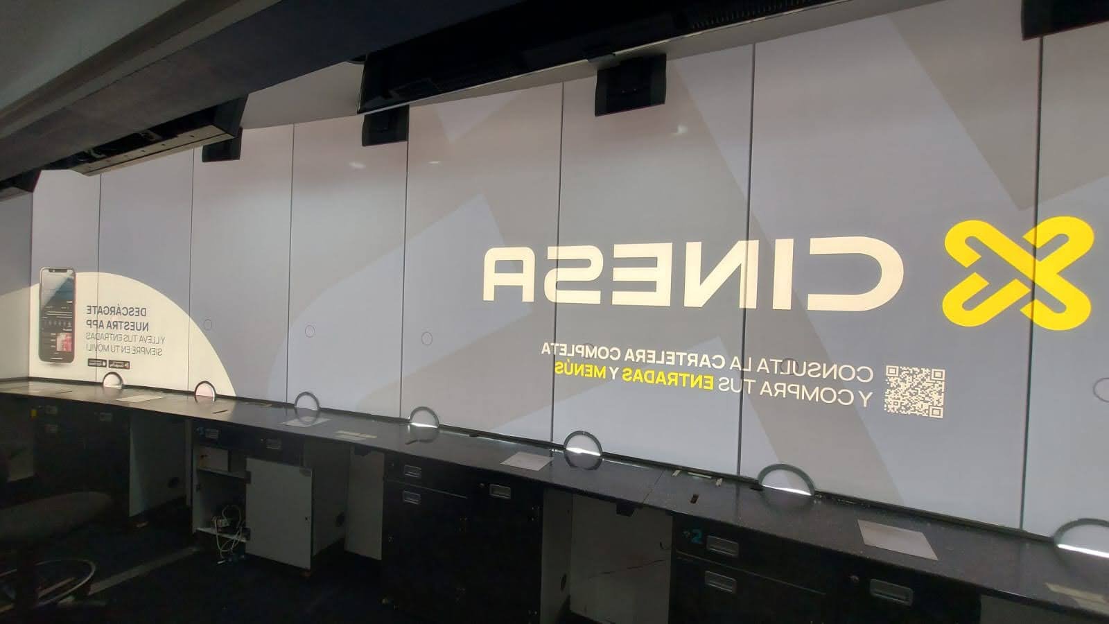 Pantallas digitales en una oficina con texto en español sobre servicios de CINECASE, incluyendo un código QR y un cartel promocional.